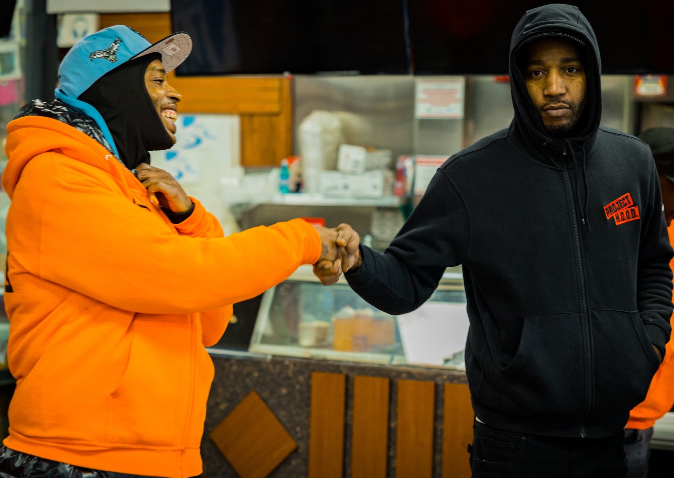 Two men shaking hands inside a store, one in an orange jacket and cap, smiling; the other in a black hoodie with an orange patch, looking serious.