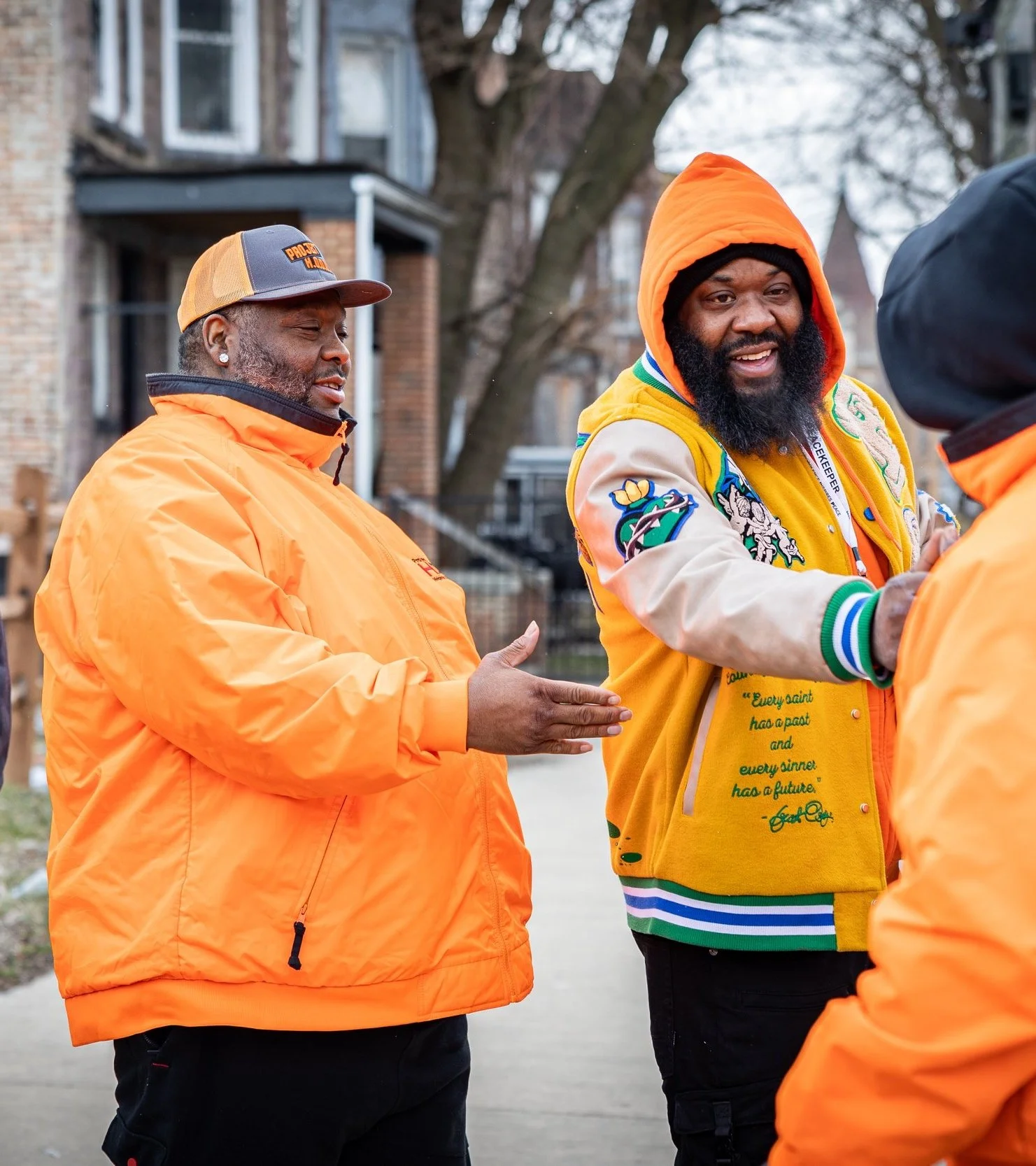 Three men shaking hands outdoors in front of residential buildings. Two men are dressed in orange jackets, and one is wearing a colorful jacket with patches, an orange hoodie, and a beard.