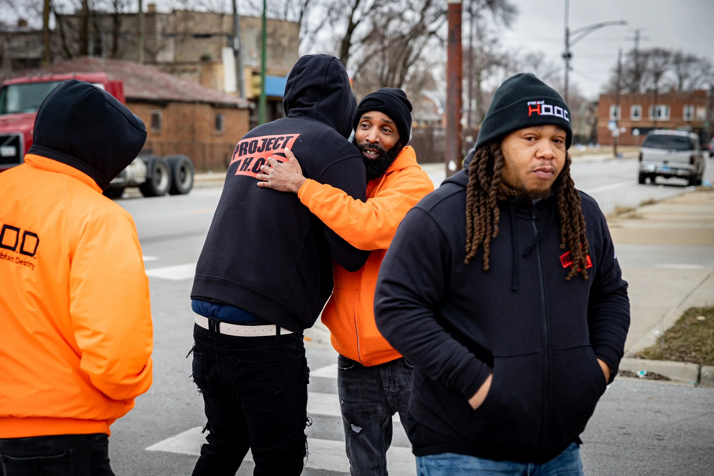 Two men in black hoodies and a man in an orange hoodie hugging each other on a street, with two other men wearing orange jackets nearby.