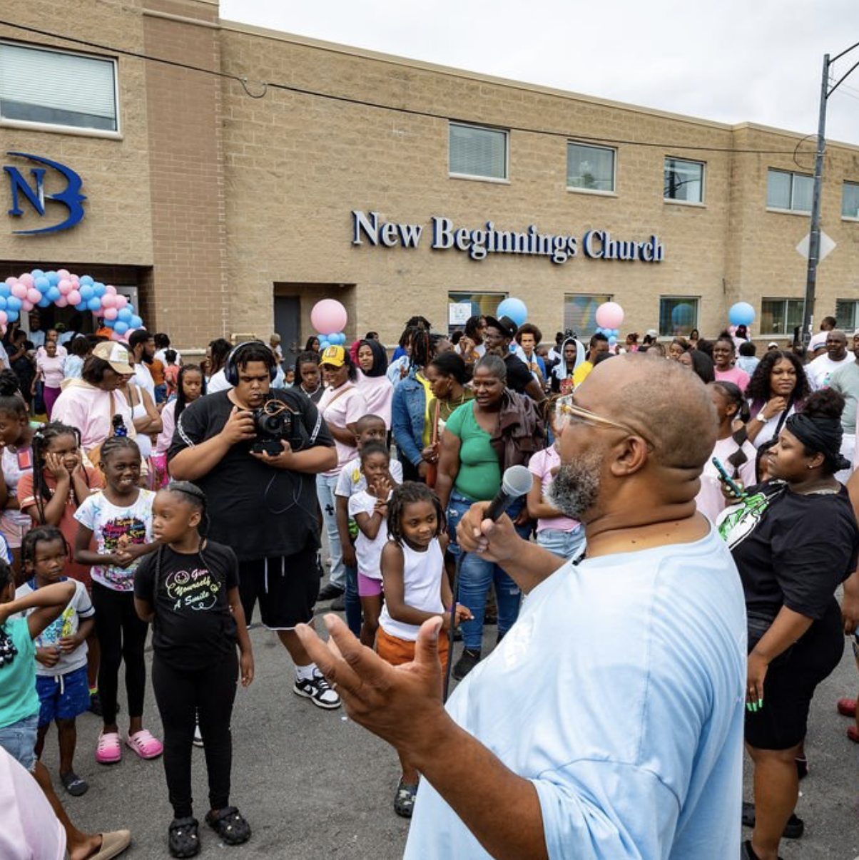 A crowd gathered outside New Beginnings Church, with balloons and decorations, as a man speaks into a microphone and a person records with a camera.