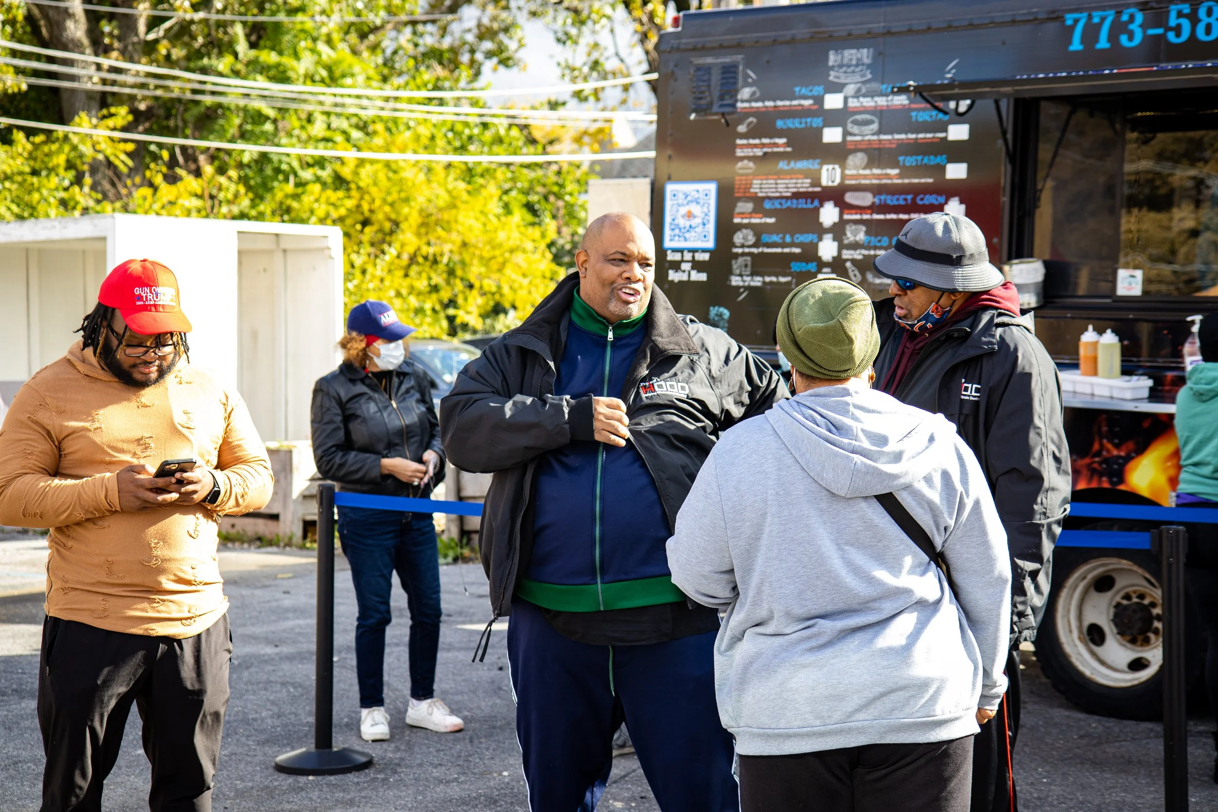 A group of people standing in front of a food truck, engaged in conversation. One person typing on a phone, others wearing jackets and masks, including a man with a beard and others in hats or caps.