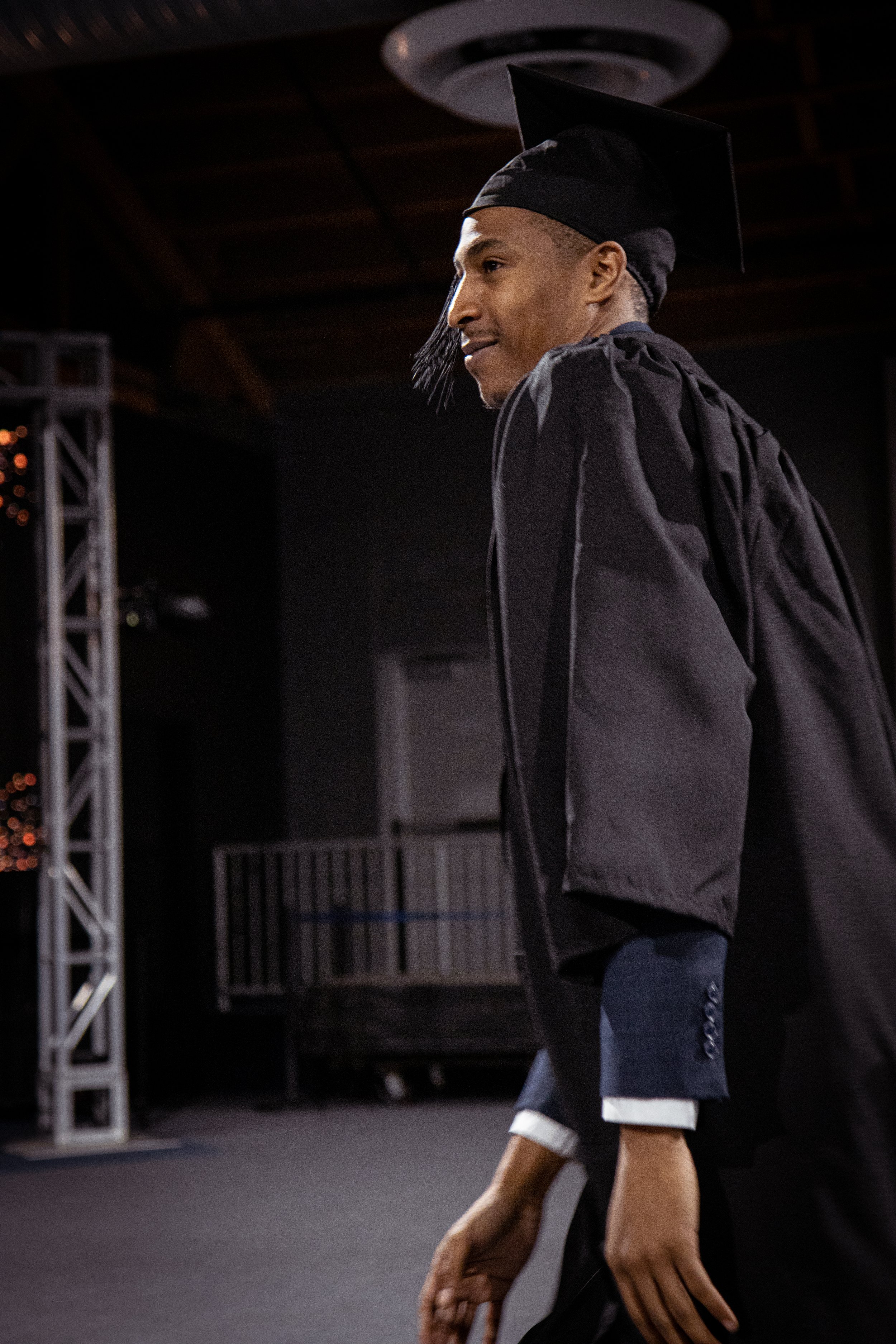 Graduate in traditional gown and cap walking indoors, smiling, with stage equipment and railing in background.