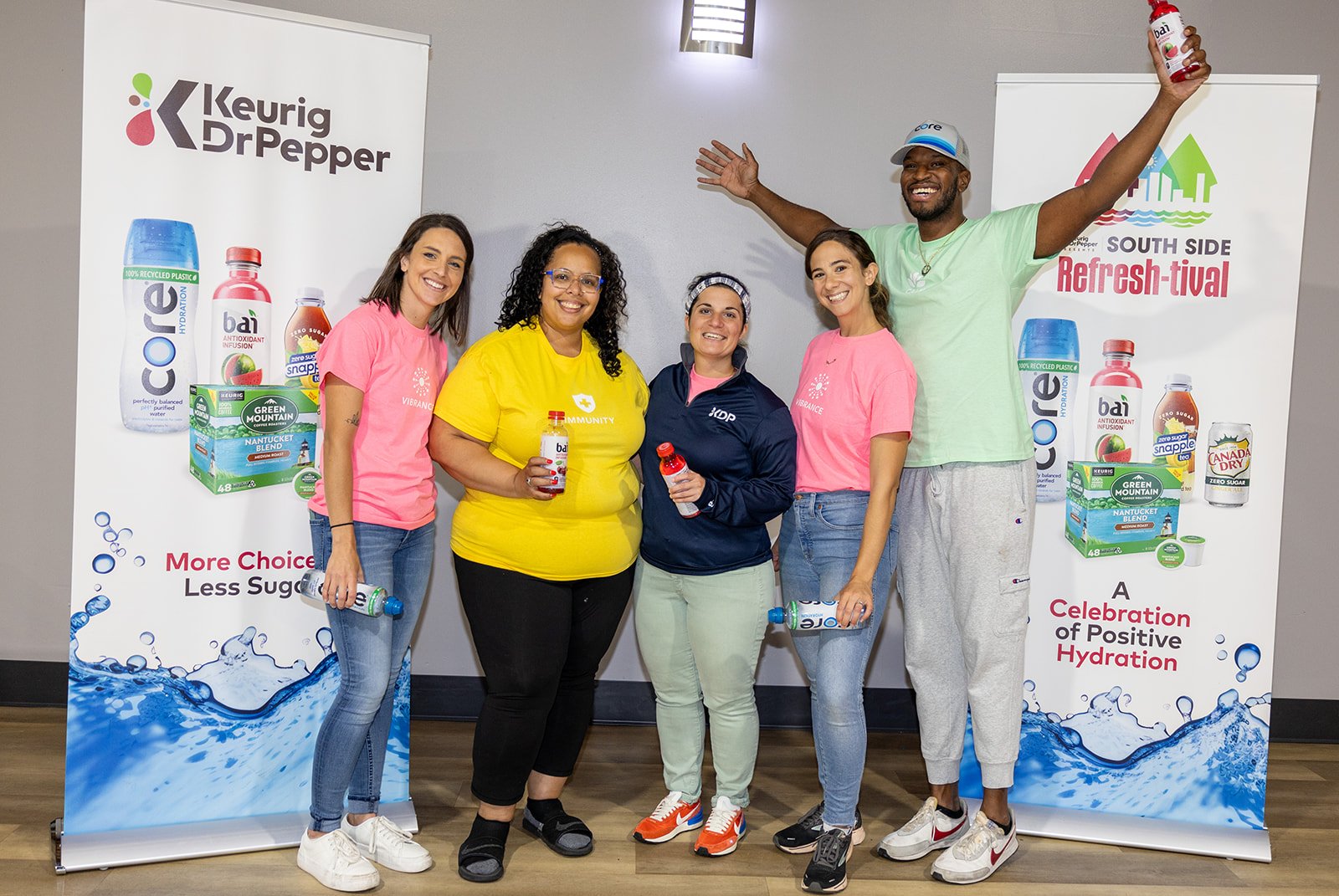 group of five smiling people standing together indoors holding various bottles of Bai beverages, with promotional banners behind them advertising Bai drinks and hydration, celebrating positive hydration and choice.