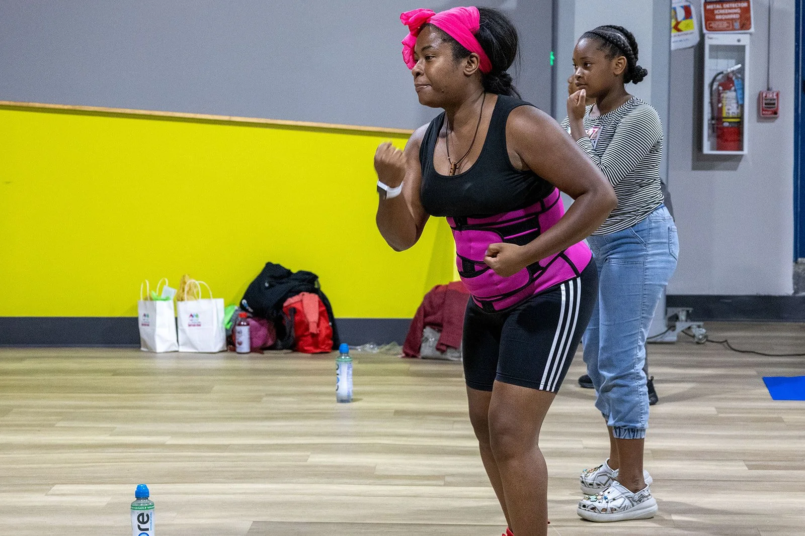 A woman wearing a black athletic outfit with a pink waist trainer and headband is exercising with a fist pump, while a young girl stands behind her watching. The scene appears to be in a gym or fitness class.