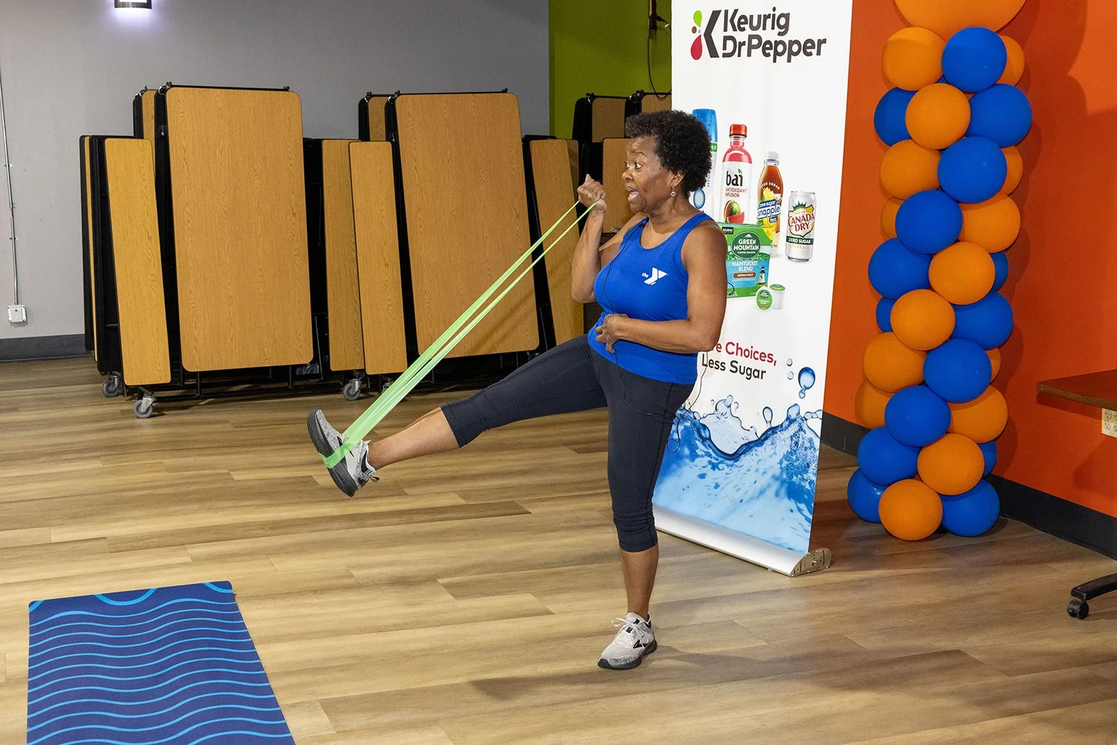 A woman in a blue tank top and black leggings is exercising with a green resistance band in a gym or fitness center. She is lifting her right leg while holding the band with her arms. There are folding tables, a Keurig Dr. Pepper sign, and blue and orange balloons in the background.