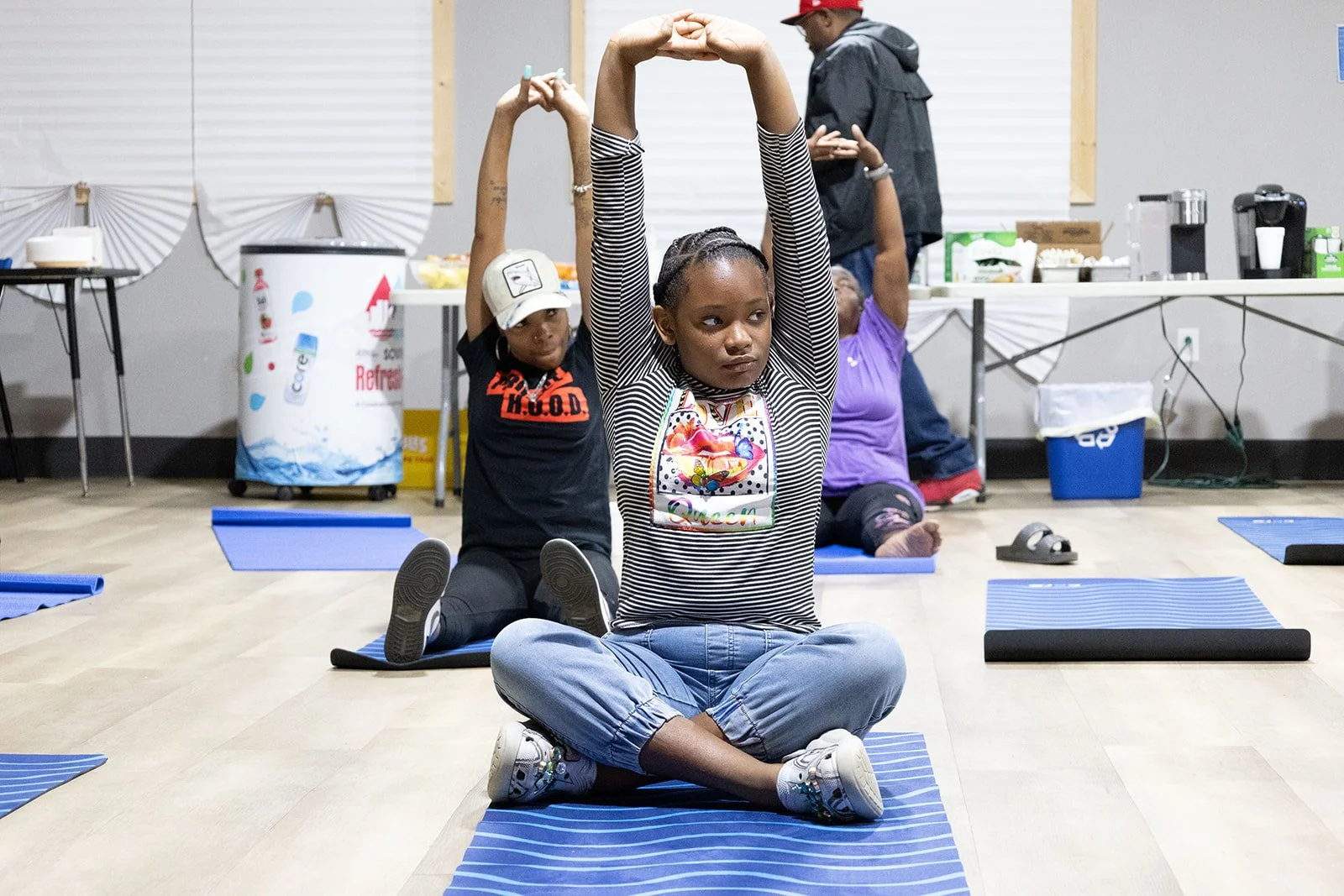 Children participating in a yoga or stretching class, sitting on yoga mats with arms raised overhead in a seated twist pose in a room with a table of refreshments in the background.