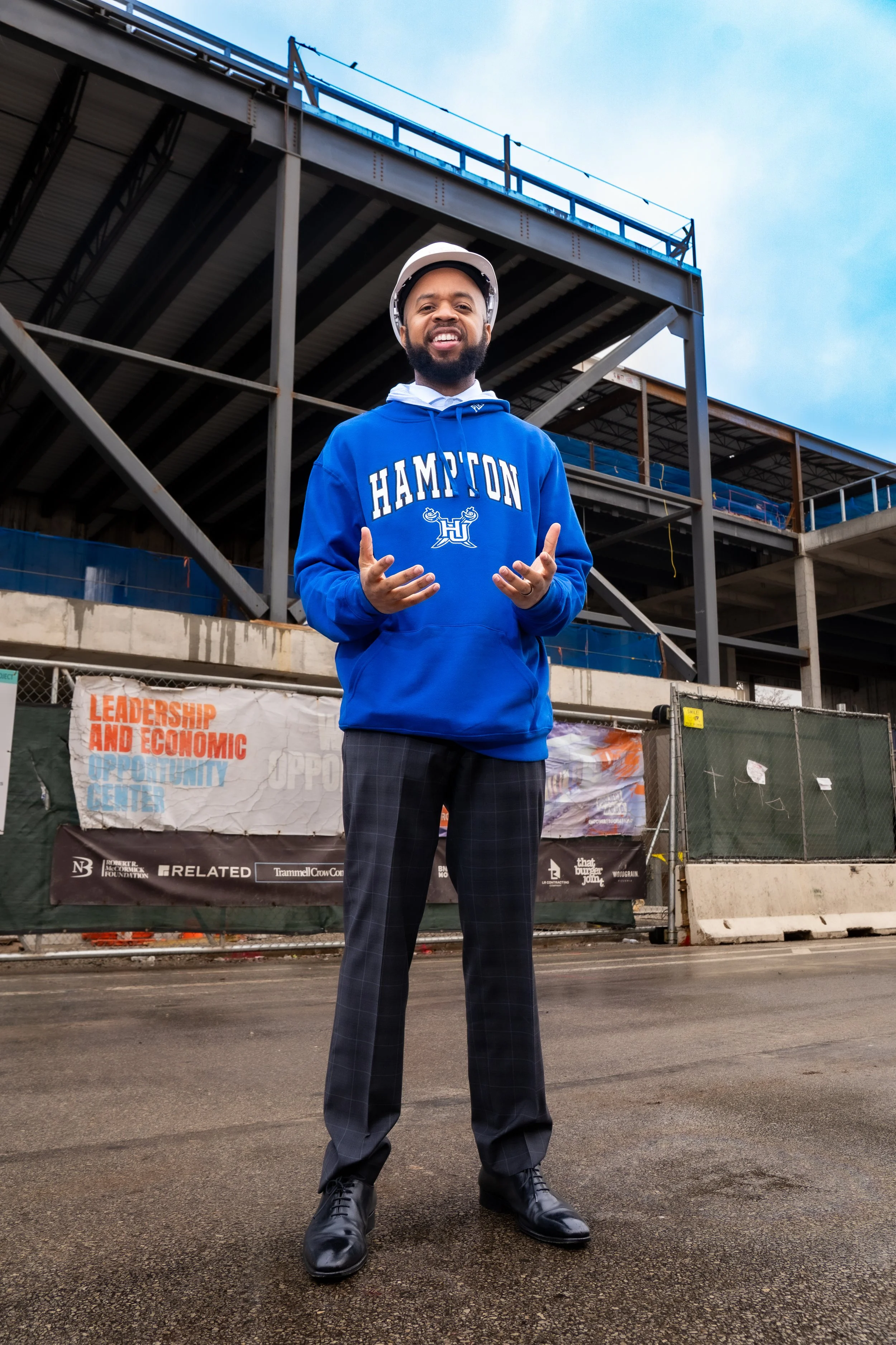 A man wearing a blue Hampton hoodie and a construction helmet standing on a construction site, smiling at the camera.
