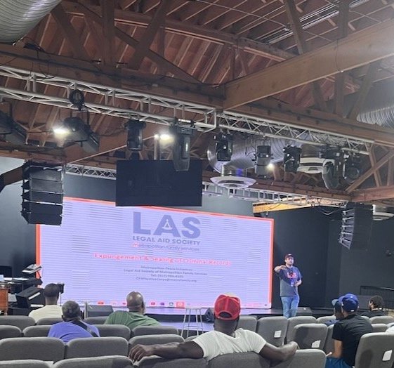 A man standing on a stage in front of a large digital screen at an indoor event. The screen displays the logo of the LAS Legal Aid Society and some text. Audience members are seated in front of the stage, some wearing hats.
