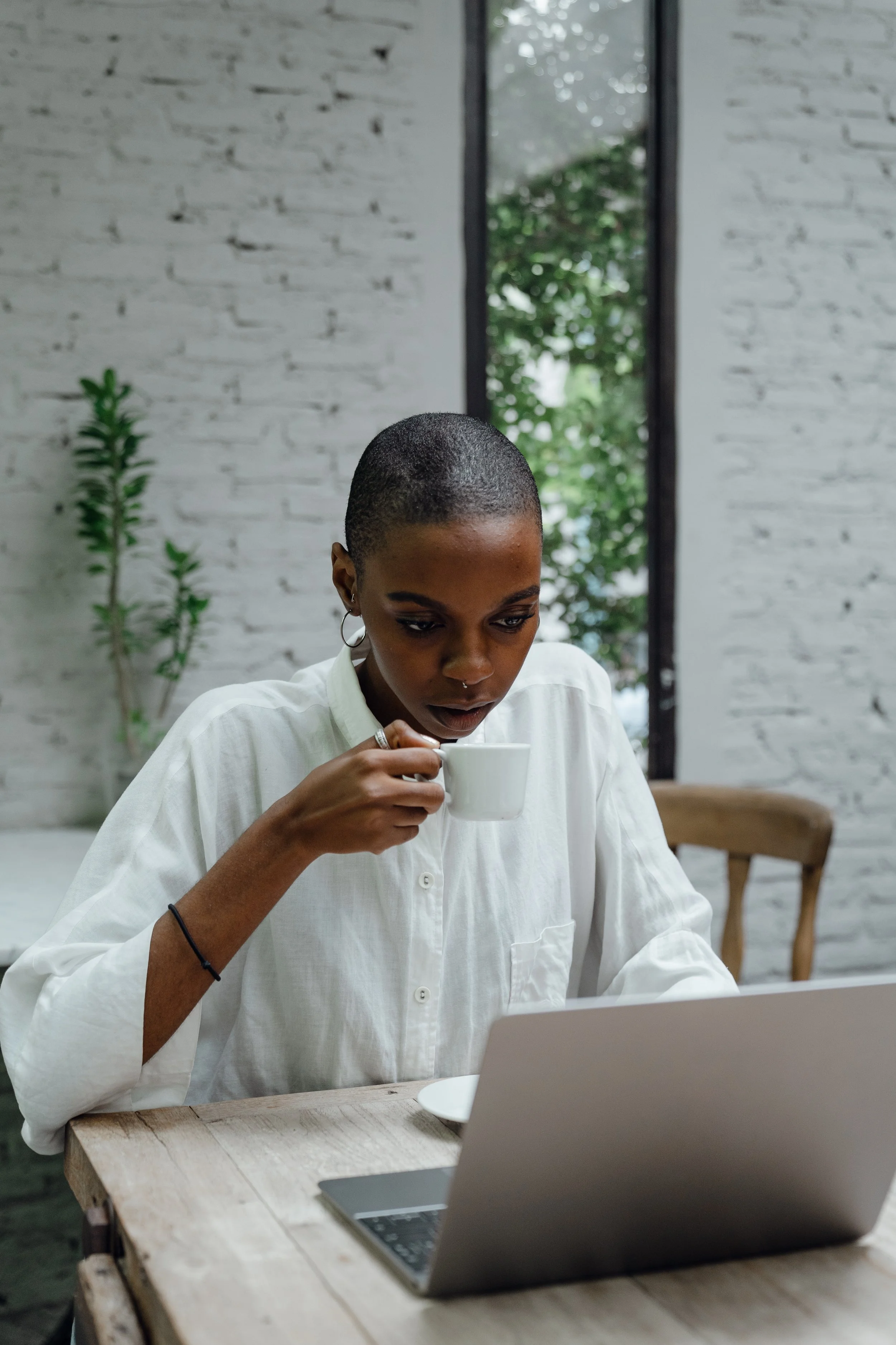A woman with a short buzz cut hairstyle wearing a white shirt, holding a coffee cup, working on a laptop at a wooden table inside a modern room with a white brick wall and a window showing greenery outside.