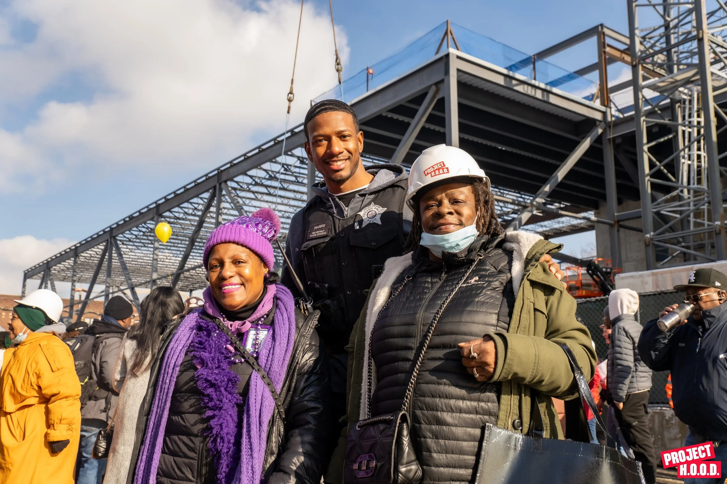 Three individuals standing outdoors at a construction site, smiling at the camera. One person in the middle is a police officer, and two women on either side are dressed warmly with purple and green jackets, scarves, and hats. The woman on the right is wearing a construction helmet and a face mask. There are other people in the background, some wearing helmets and jackets. A building under construction is visible behind them.