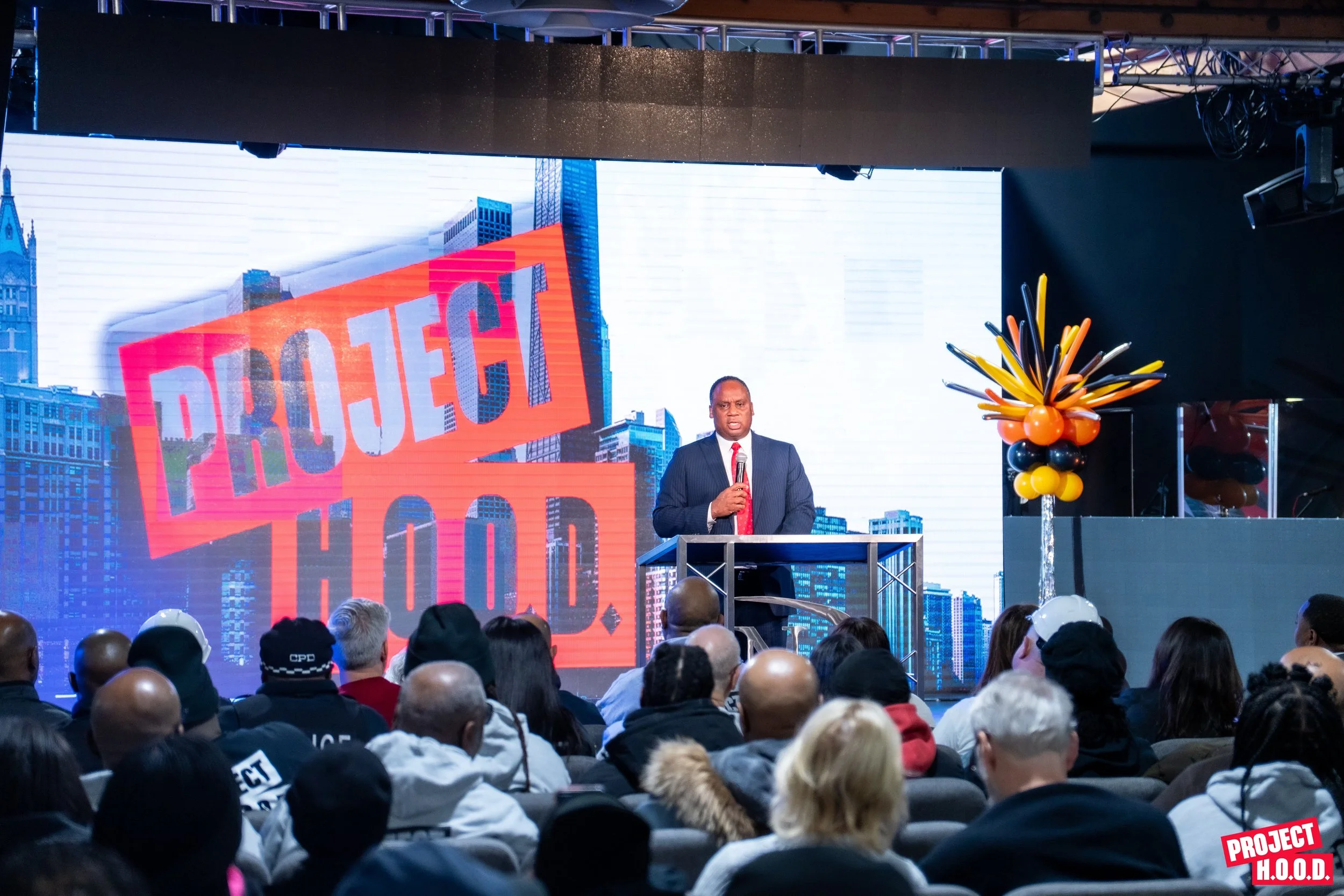 A man in a suit and red tie speaking at a podium during a conference, with a large digital screen behind him displaying the words 'PROJECT H.O.O.D.' and a cityscape background. Audience members seated in front are watching.