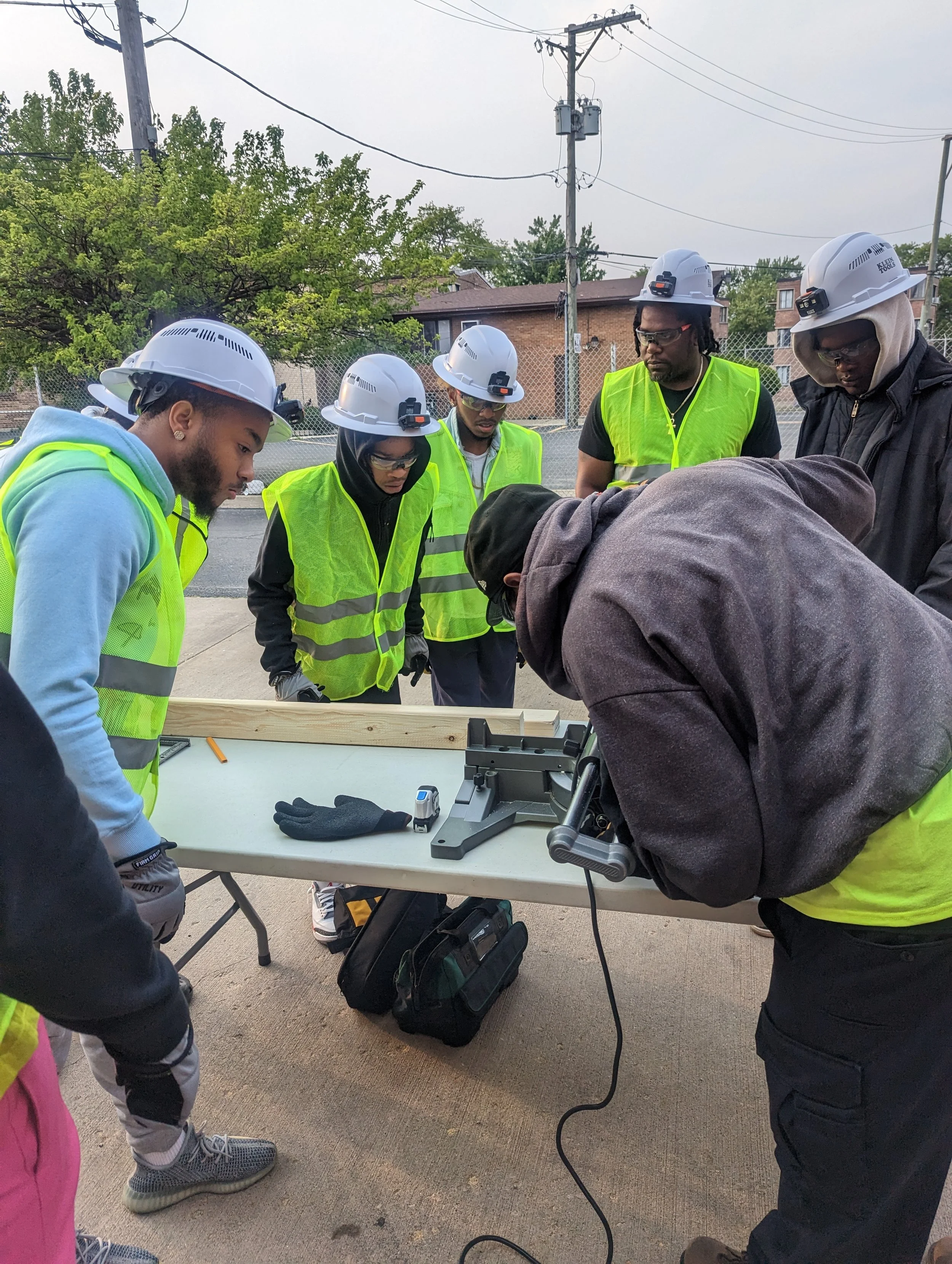 Group of construction workers in safety gear, including helmets and vests, gathered around a work table outdoors, likely receiving instructions or training on power tools and construction safety.