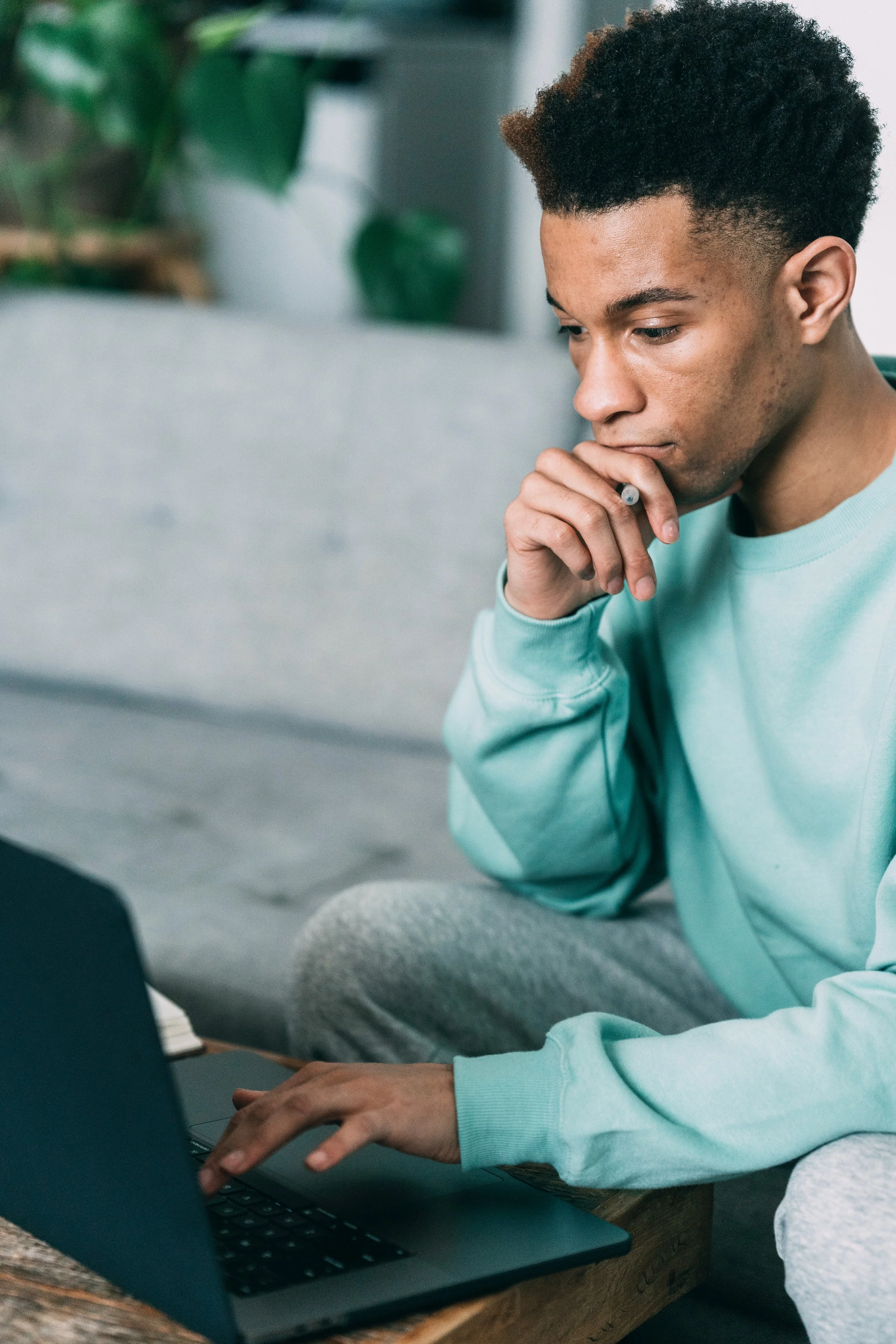 A young man with curly hair sitting on a couch, looking at his laptop with a thoughtful expression, holding a pen near his mouth.
