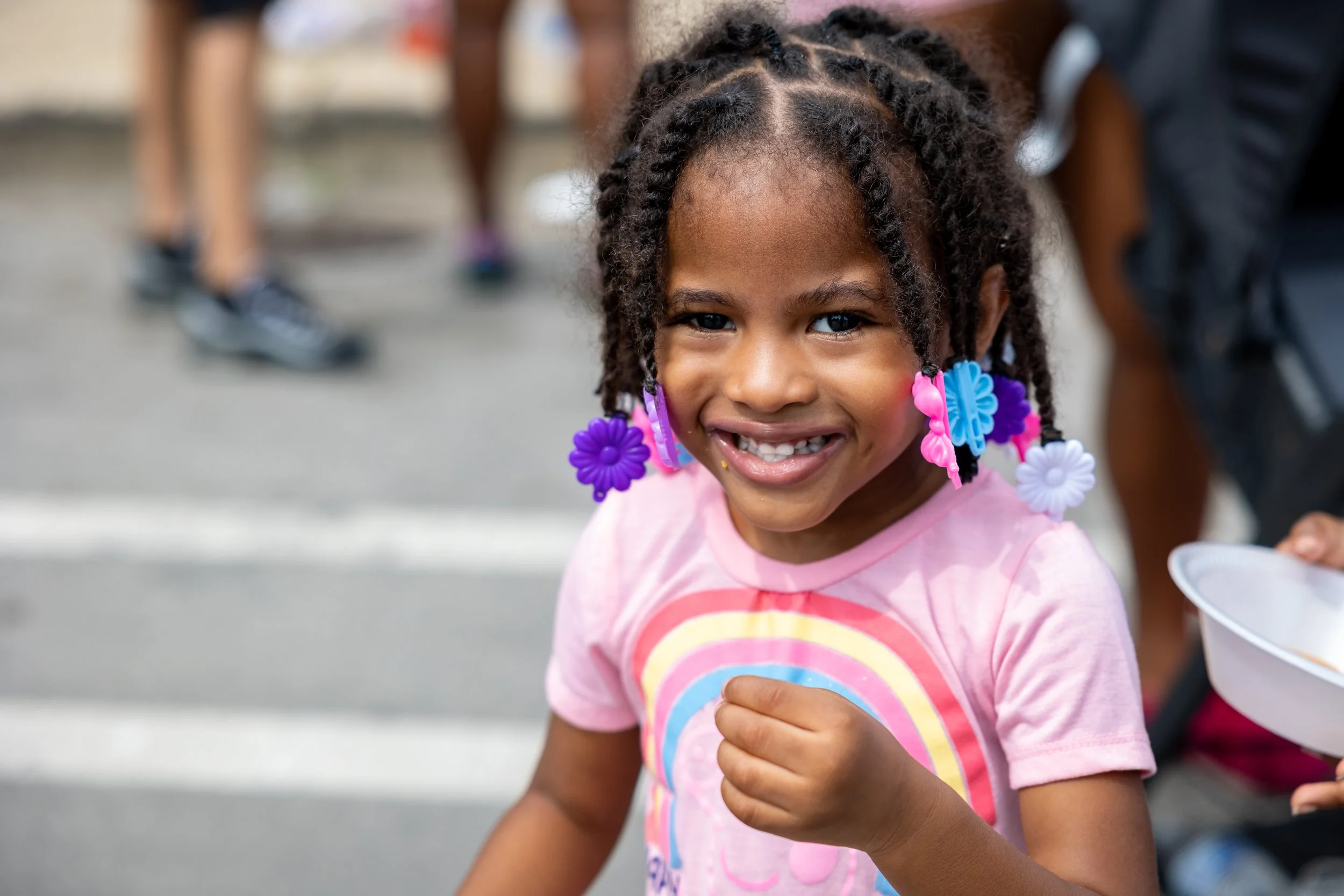 A young girl with braided hair decorated with colorful flower beads, smiling and showing her teeth, wearing a pink rainbow t-shirt, standing on the street during daytime.