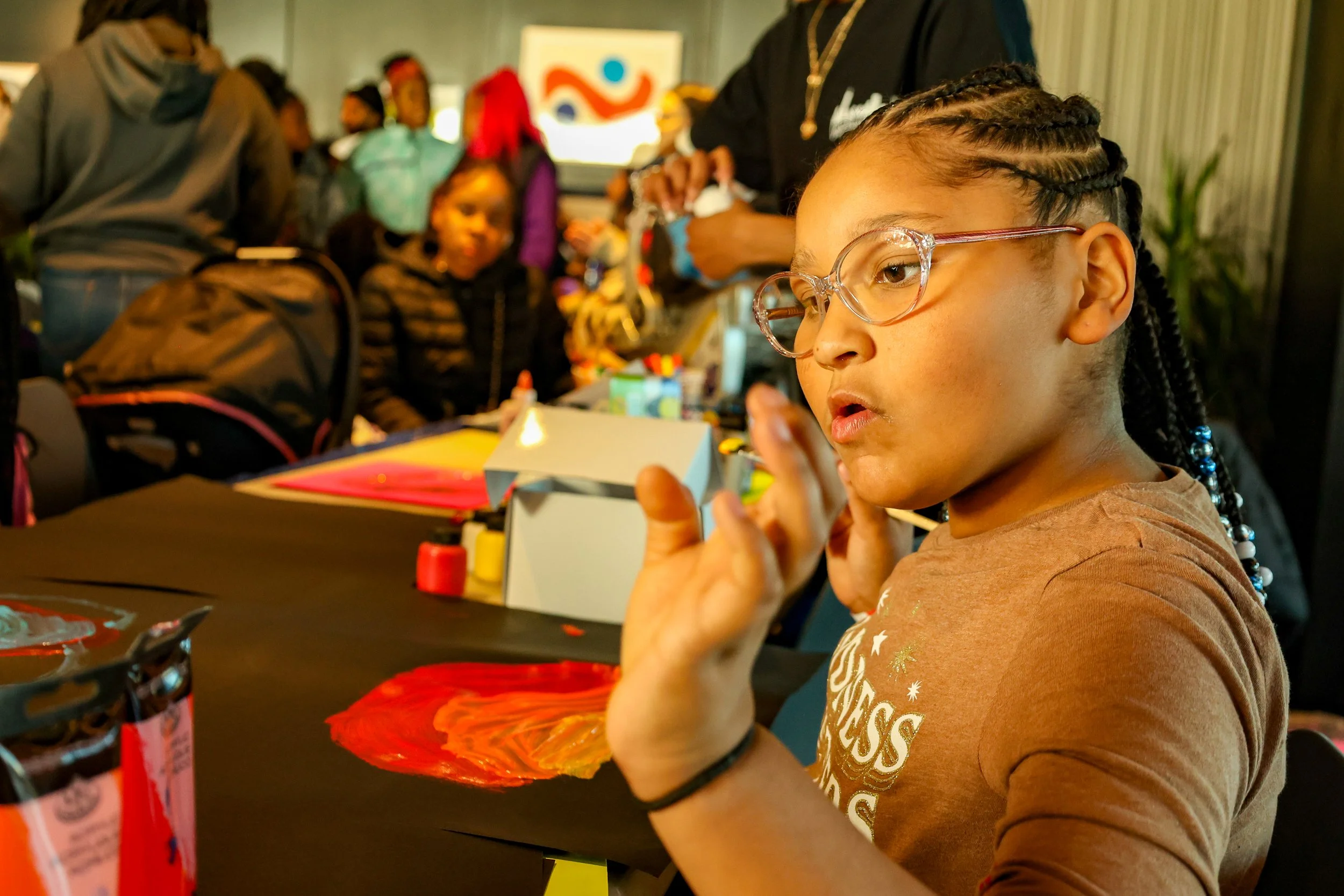 A young girl with glasses and braids holds a paintbrush at an arts and crafts activity, surrounded by children and adults in a busy indoor setting.