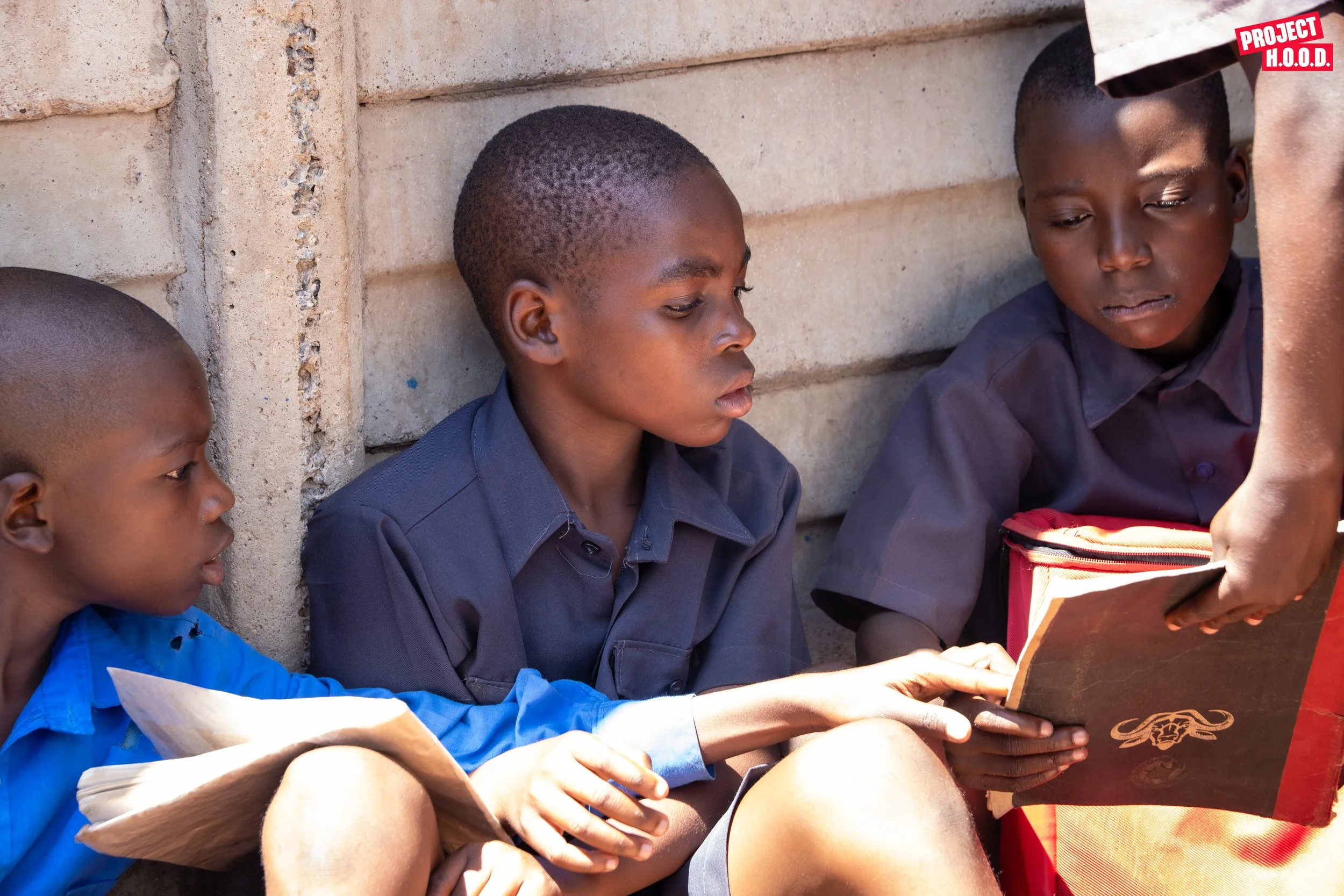 Three children in school uniforms sitting against a concrete wall, reading books, with an adult handing them a book.