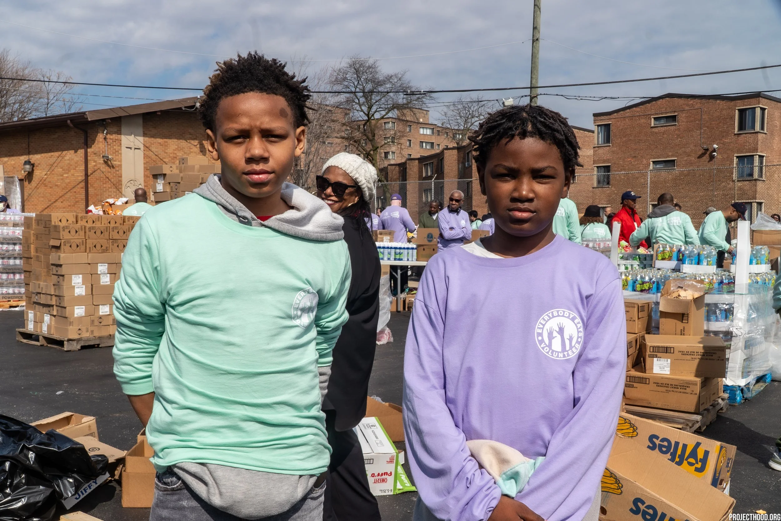 Two young boys standing outside at a community event, with tables of supplies and people in the background, on a partly cloudy day.