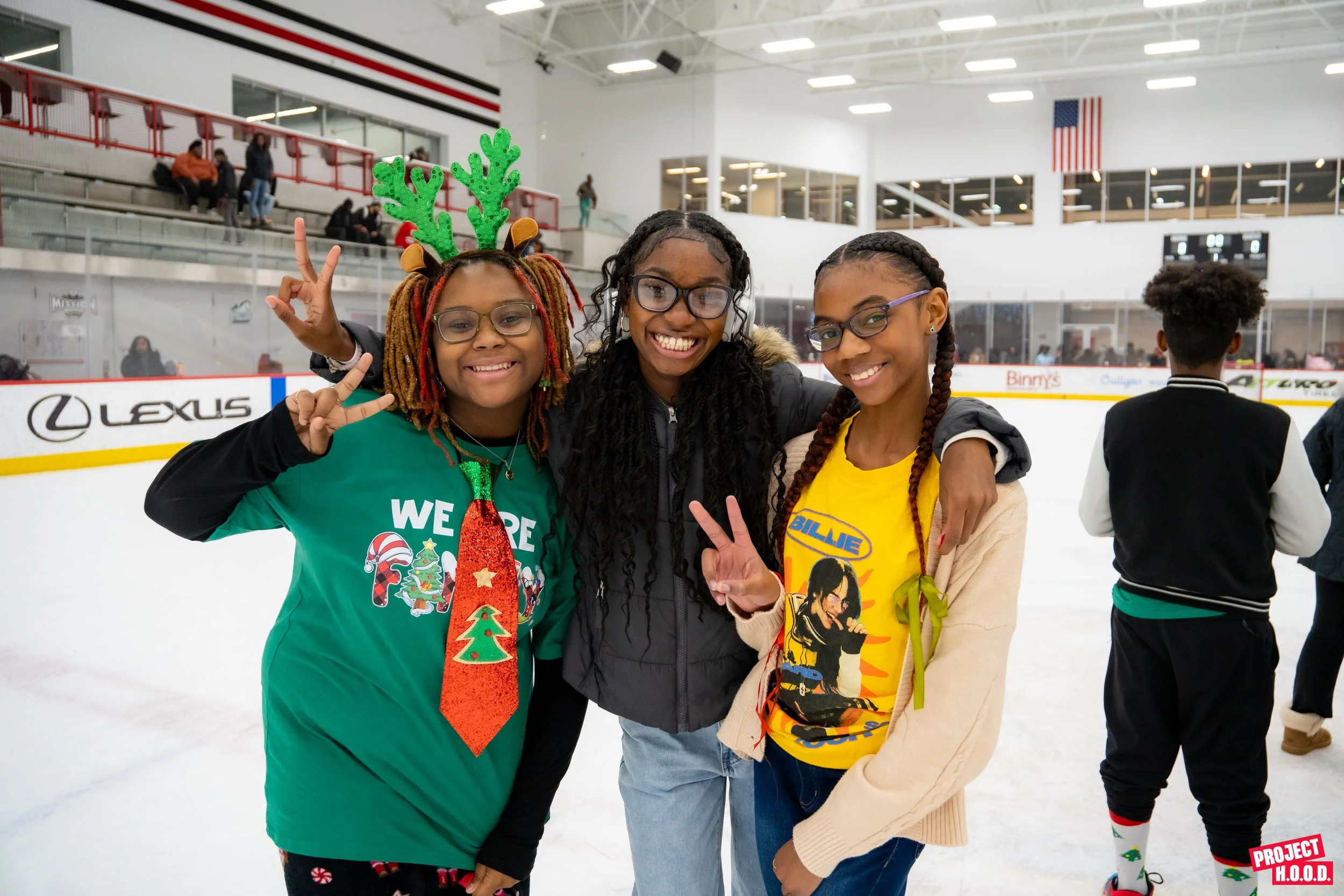 Three young girls smiling and making peace signs on an ice rink, with holiday-themed clothing and accessories, and other skaters in the background.