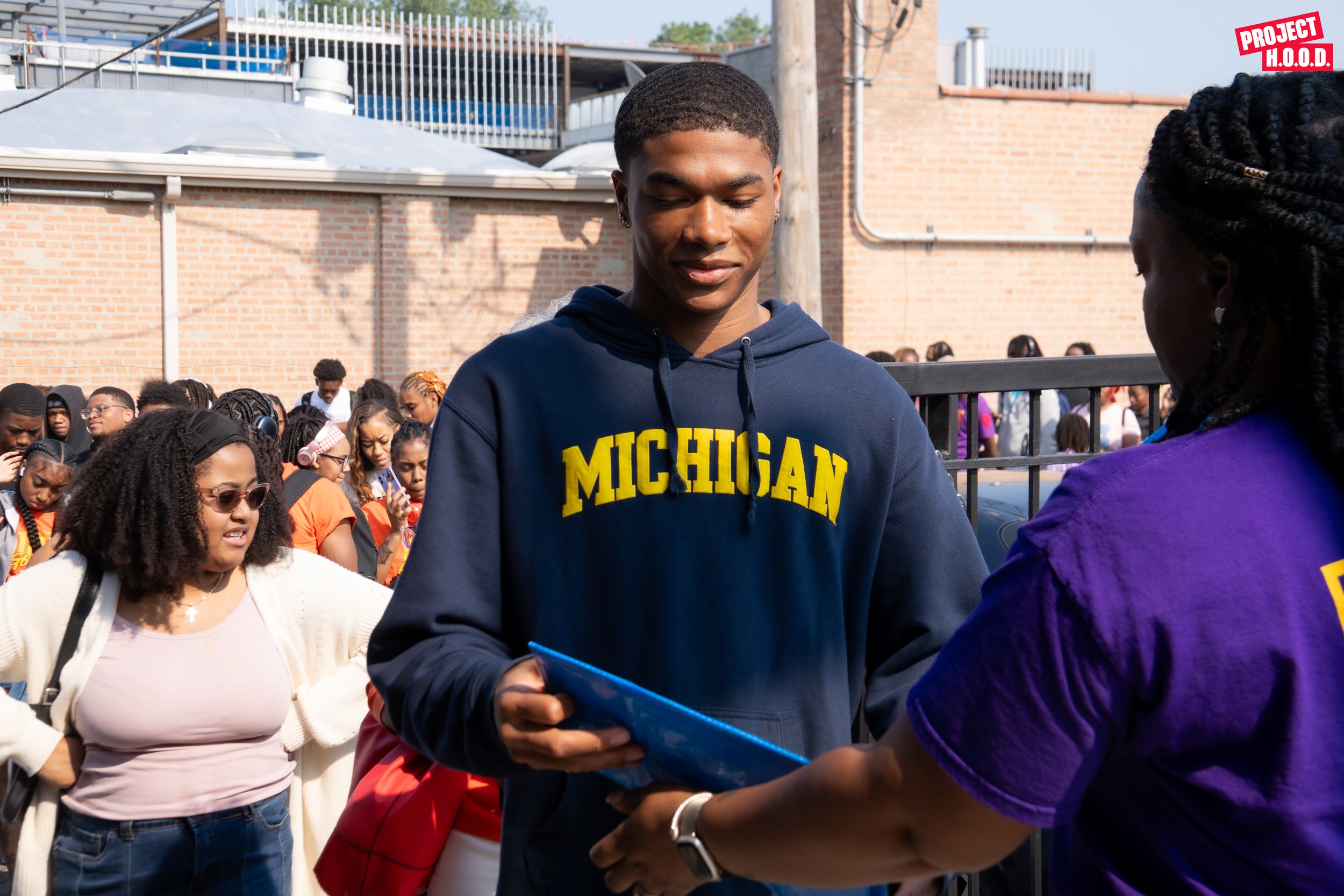 A young man wearing a Michigan hoodie is receiving a document or certificate from a woman in a purple shirt outdoors during a crowd event.