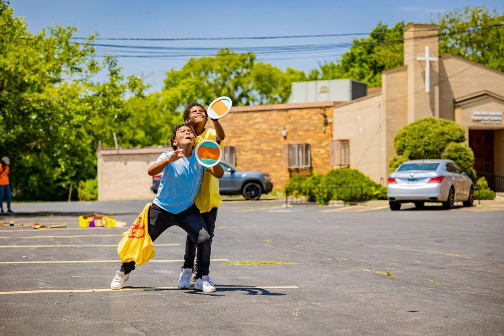 Two children playing outside in an empty parking lot, throwing frisbees, with a church and cars in the background on a sunny day.