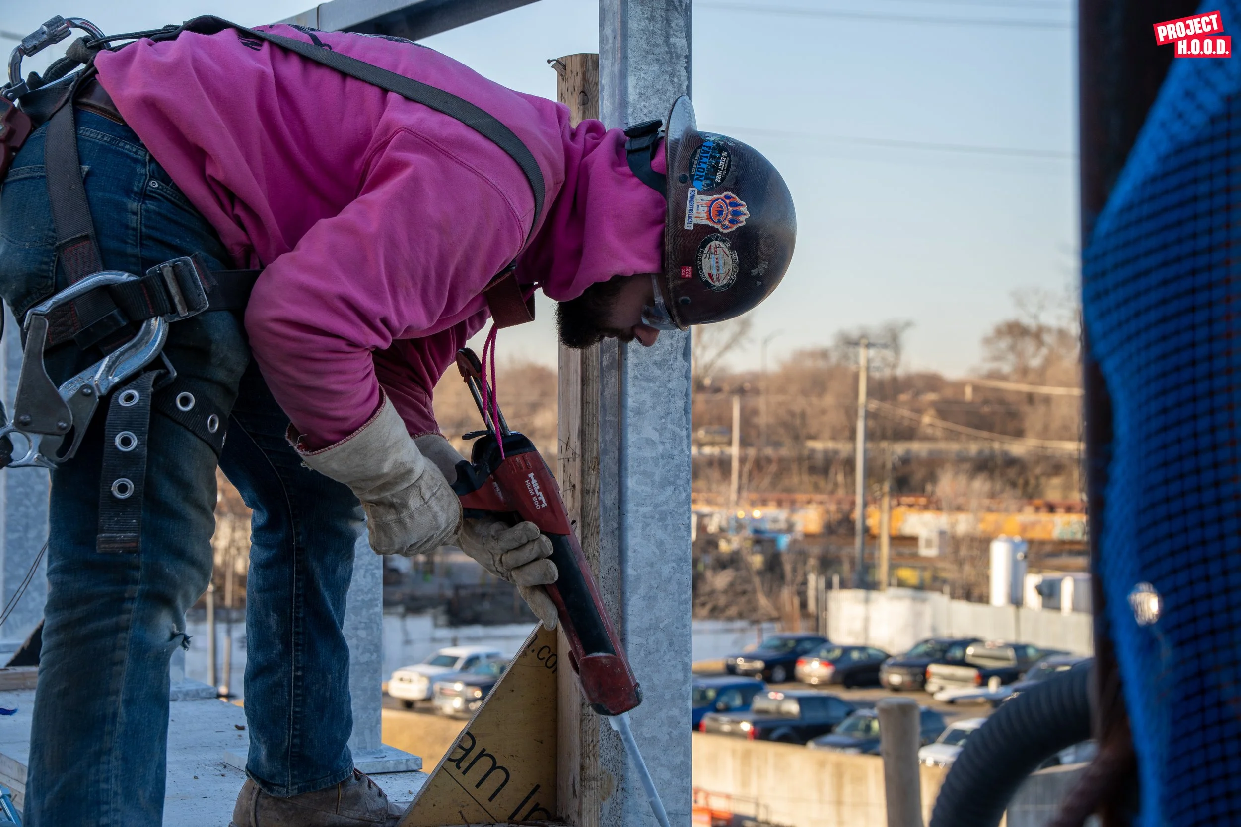 A construction worker wearing a pink hoodie, gloves, and a safety helmet is using a power drill to secure a metal pole at a construction site with a parking lot and buildings in the background.