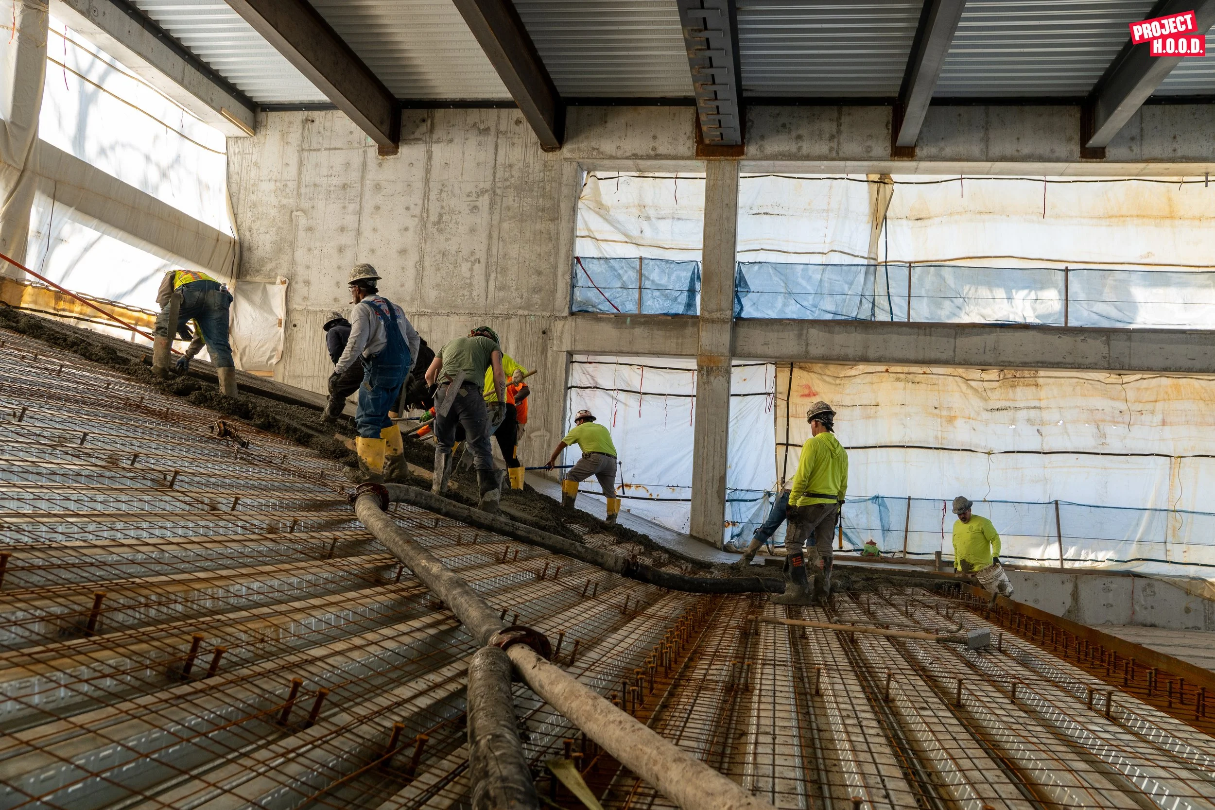 Construction workers pouring and leveling concrete on a building floor with rebar reinforcement, inside a partially enclosed structure with scaffolding and plastic sheeting.