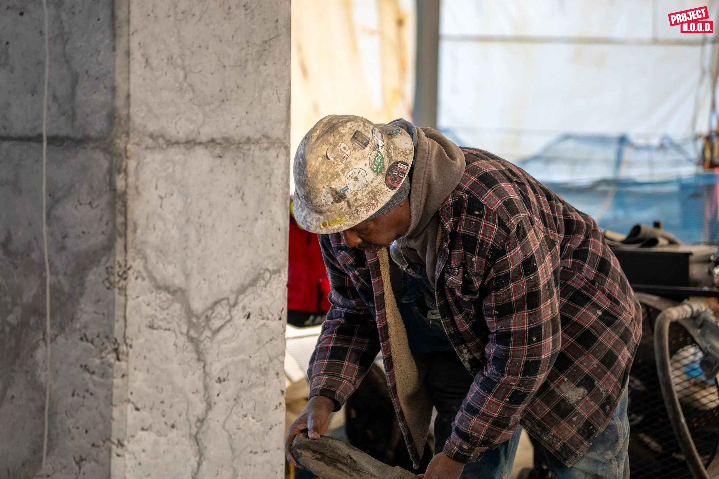 A construction worker wearing a dusty hard hat and plaid jacket is working on a concrete wall in an indoor construction site.