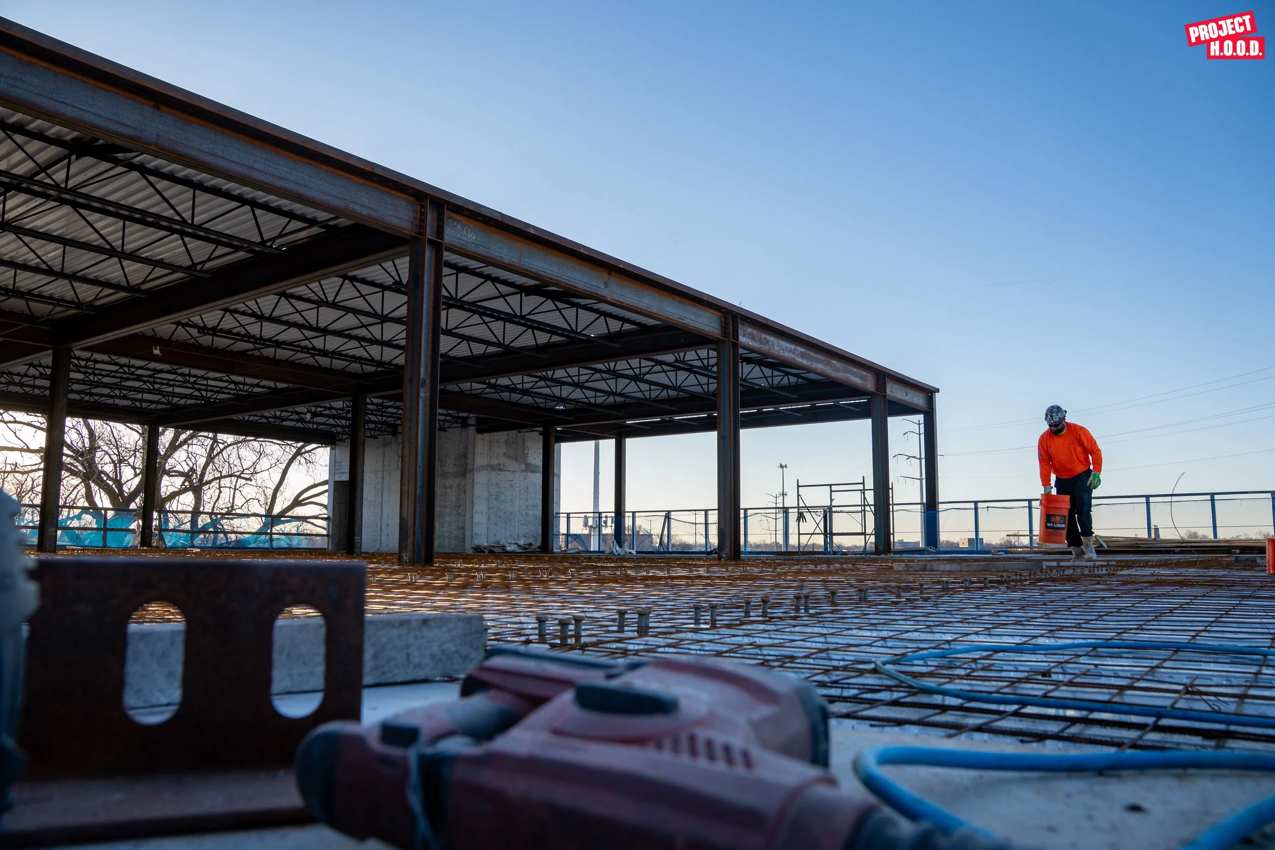 A construction worker walking on a building under construction, holding an orange bucket. The building has a metal framework and rebar on the floor. The scene is outdoors with a clear blue sky.