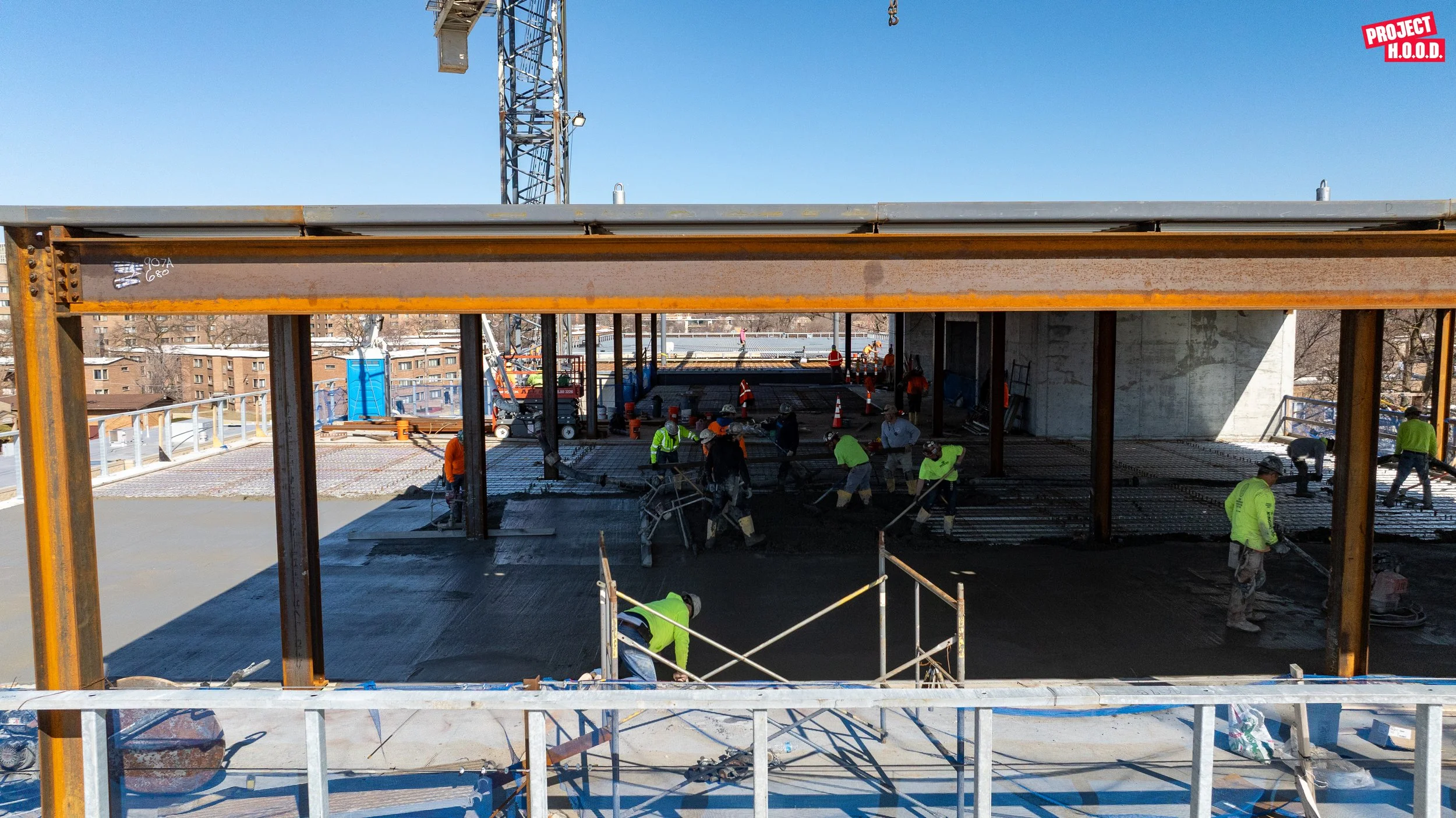Construction workers in bright yellow and orange safety vests pour concrete on a building floor under a blue sky, with a large crane and residential buildings in the background. A red and white logo reading 'PROJECT H.O.O.D.' is in the top right corn