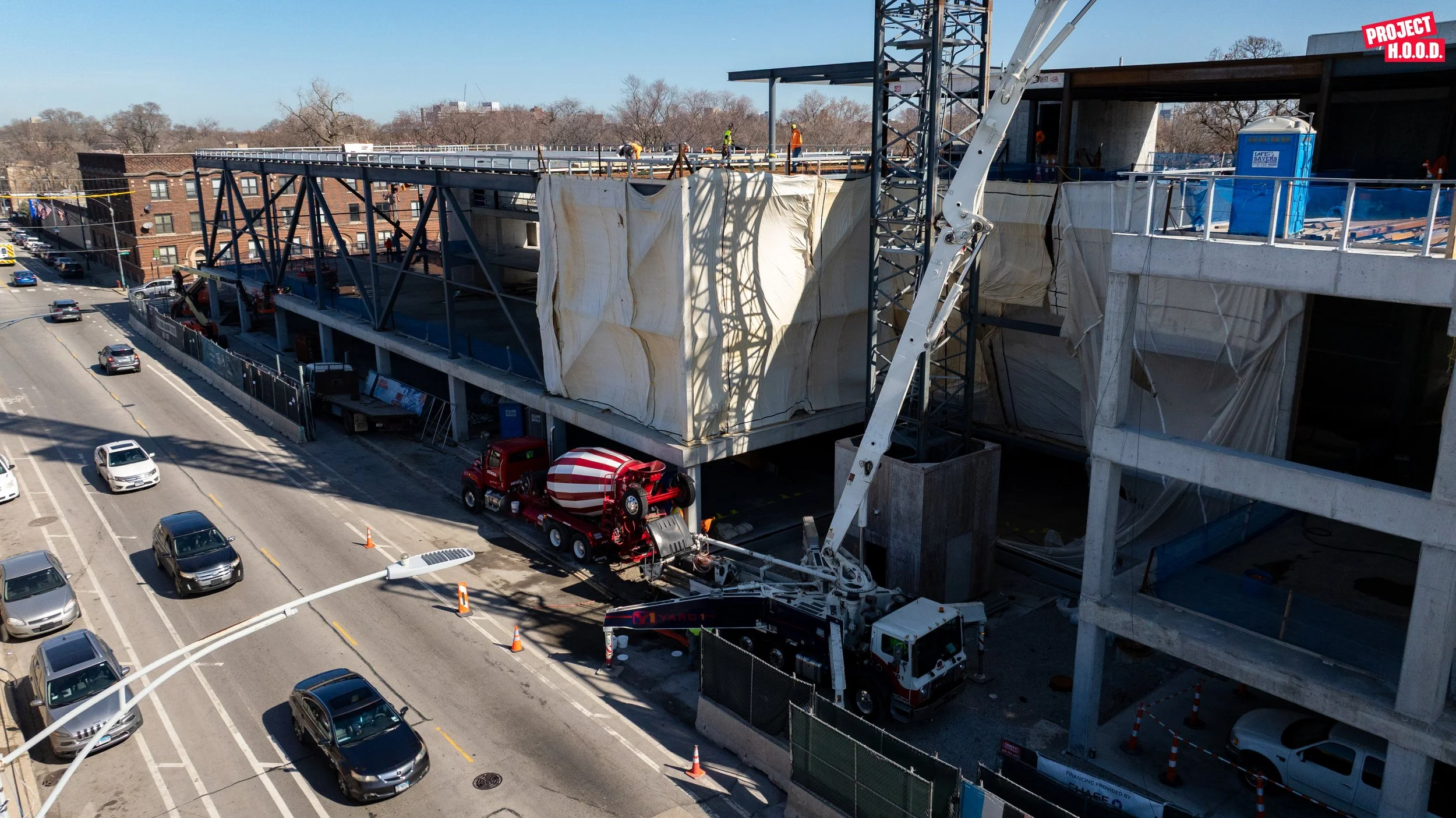 Construction site on a multi-story building with steel framework, construction workers, and a cement mixer truck on the street below.