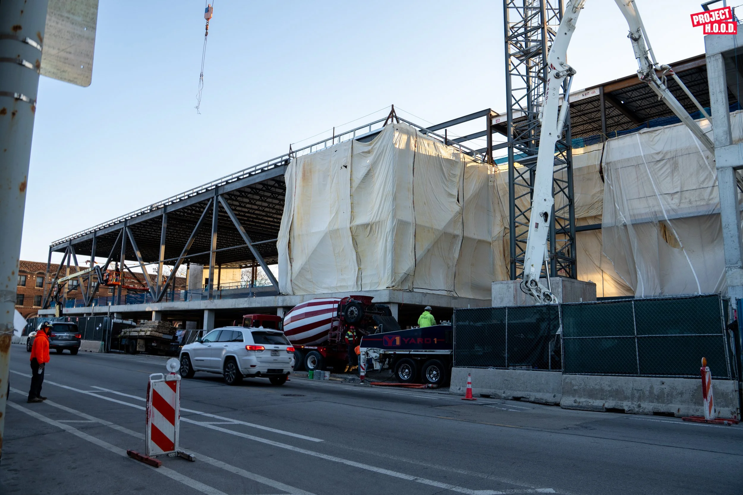 Construction site with partially built structure covered by plastic tarps, construction crane, and vehicles including a cement mixer truck, on a city street with traffic cones and workers in safety gear.
