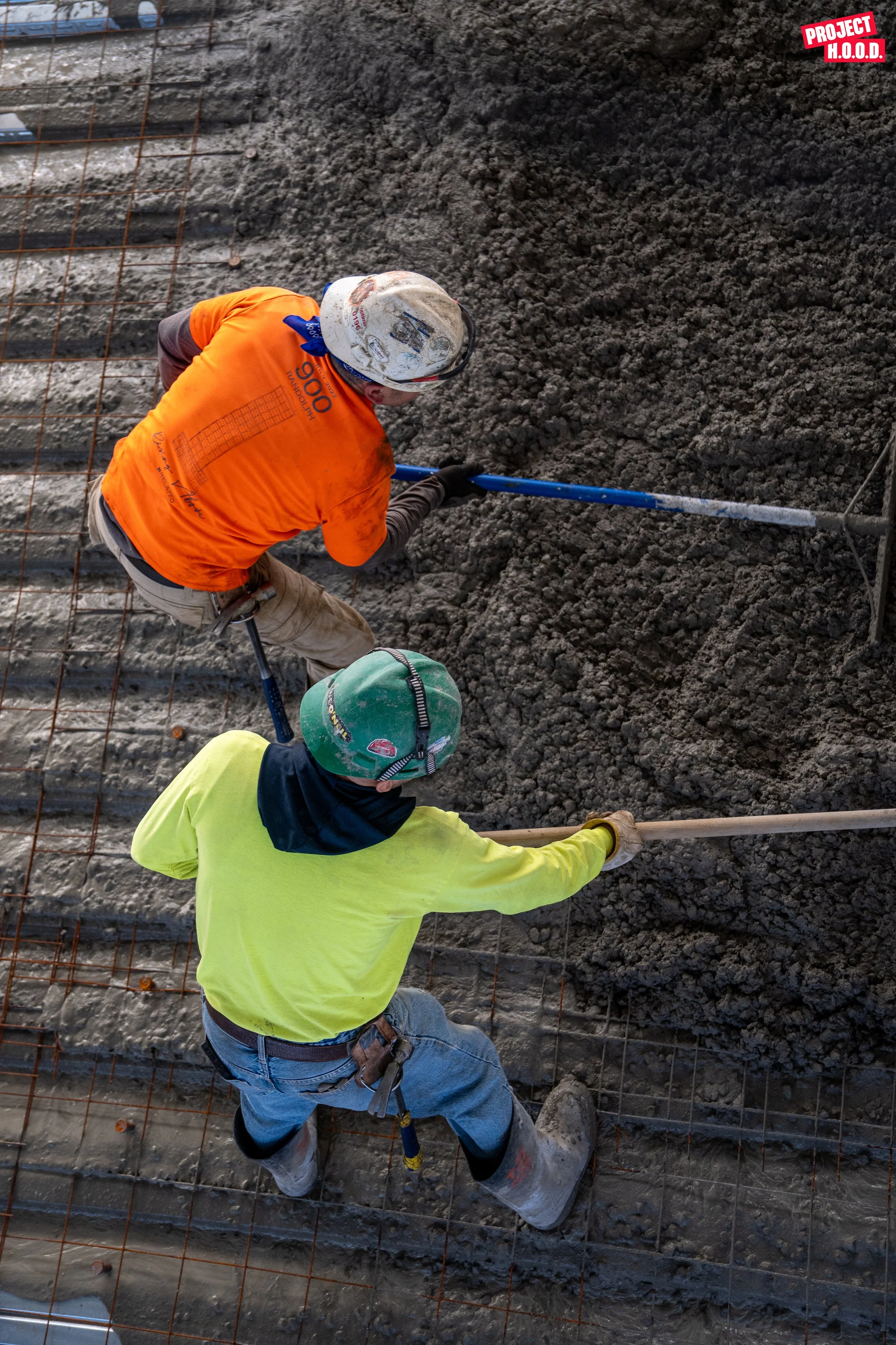 Two construction workers in helmets and safety gear spreading concrete on a rebar steel mesh at a construction site.