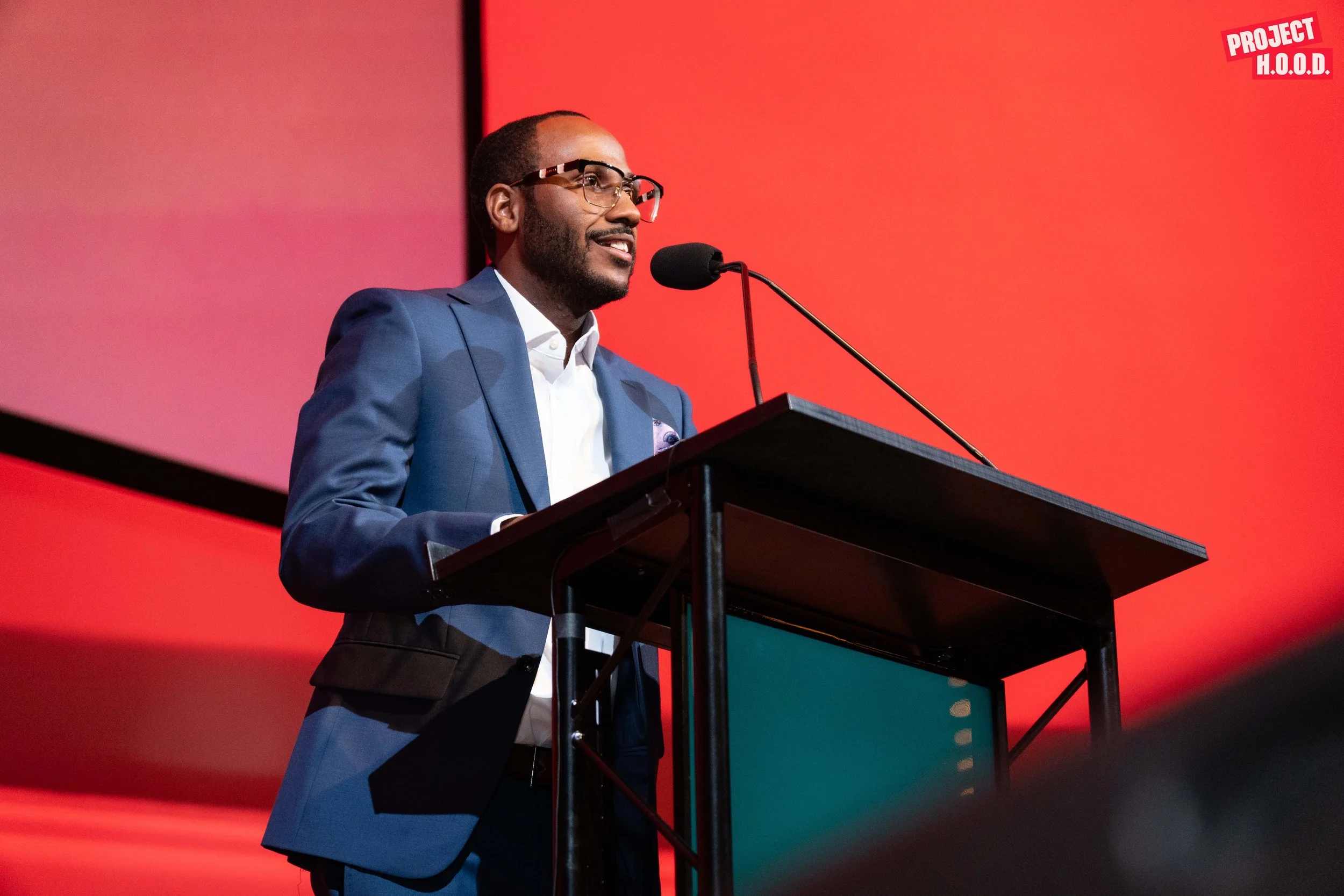 Man in blue suit and glasses speaking at a podium with a microphone, against a red background with Project H.O.O.D. logo.