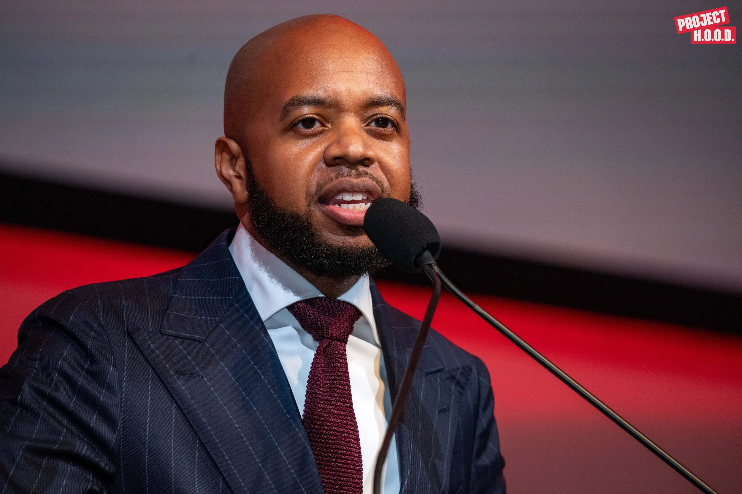 A man in a navy pinstripe suit, white shirt, and burgundy tie speaking into a microphone at a conference or event, with a red and black background. The top right corner features the logo for Project H.O.O.D.