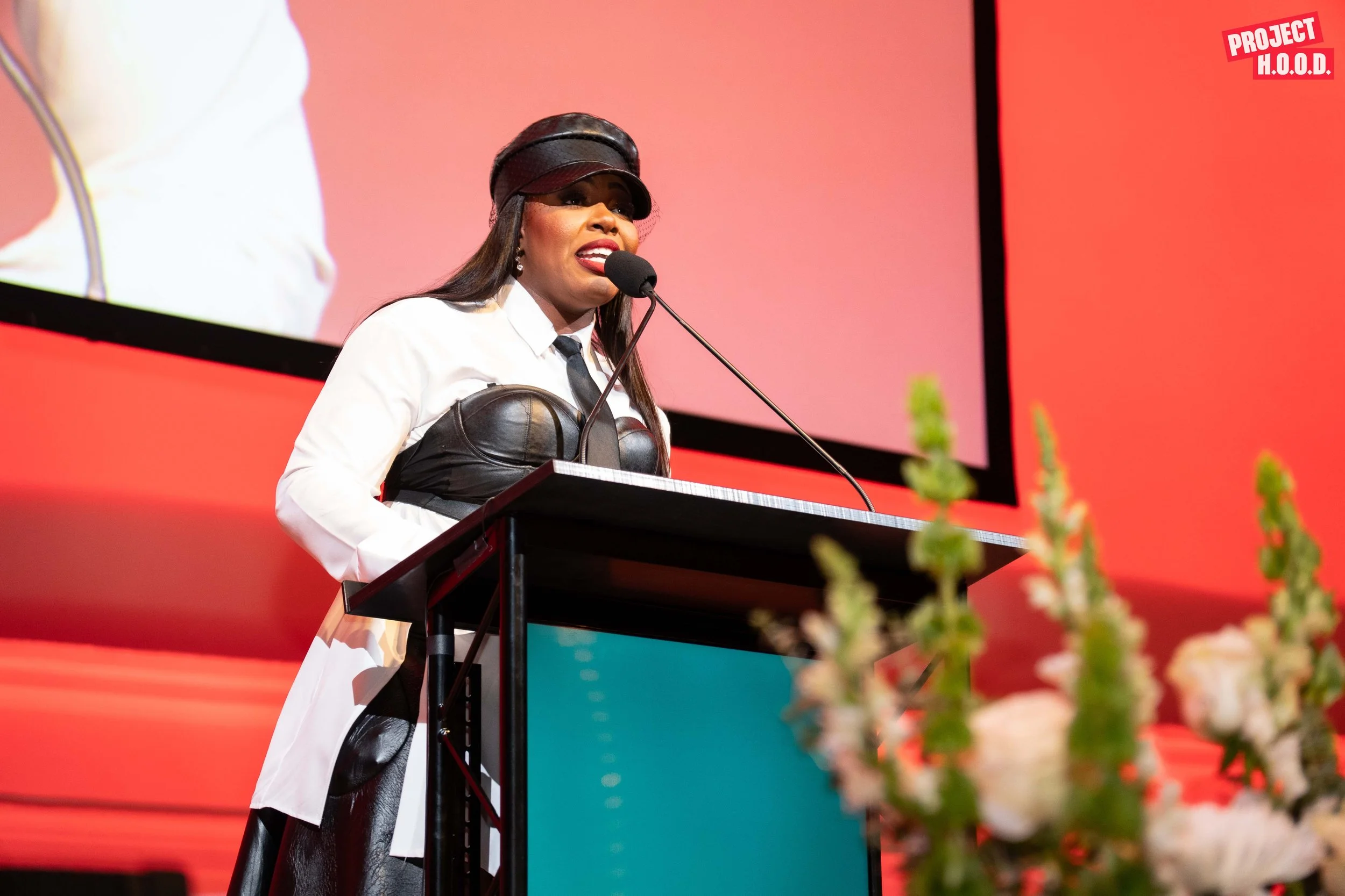 A woman wearing a white shirt, black leather corset, black leather hat, and tie speaking at a podium with a microphone at a conference or event, with a large screen in the background displaying a close-up of her and a floral arrangement in the foregr