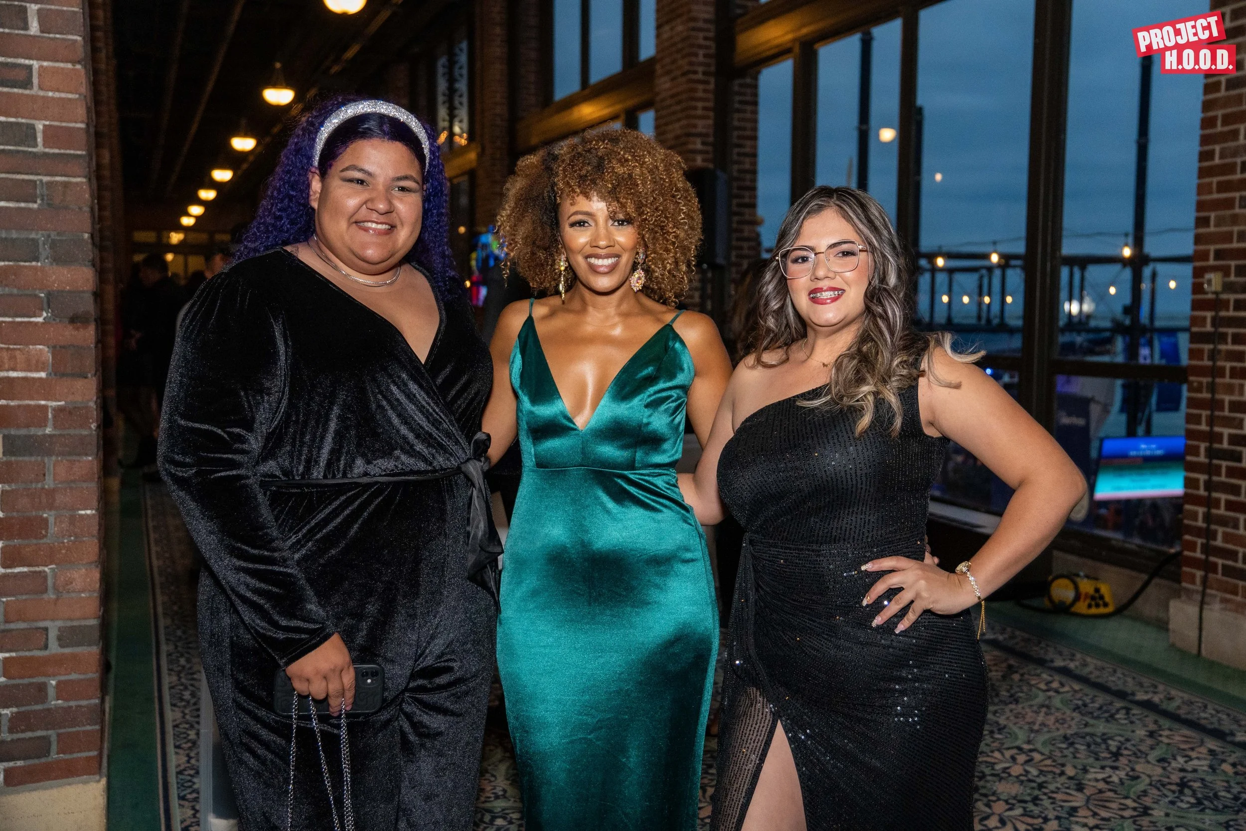 Three women dressed in glamorous evening gowns standing together indoors, with a brick wall on the left and large windows behind them showing a cityscape at dusk. The woman on the left wears a black velvet dress, the woman in the middle wears a teal 