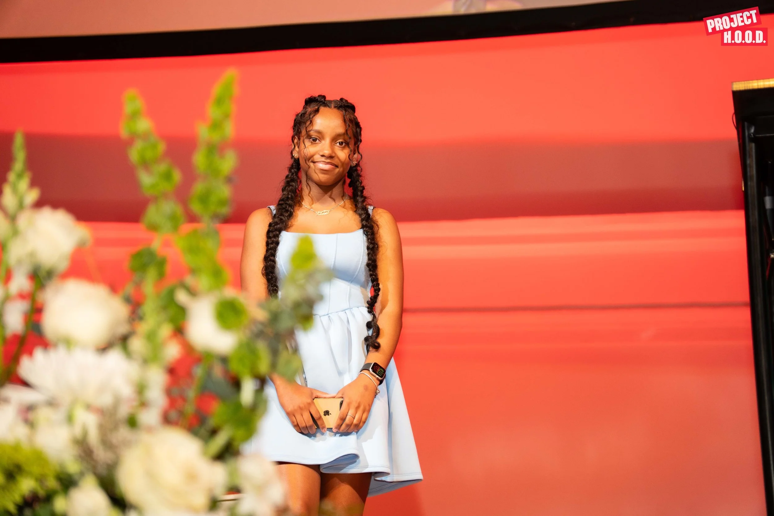 A young woman with long braided hair, wearing a white dress, standing in front of a red background at an event. She is holding a smartphone and smiling.
