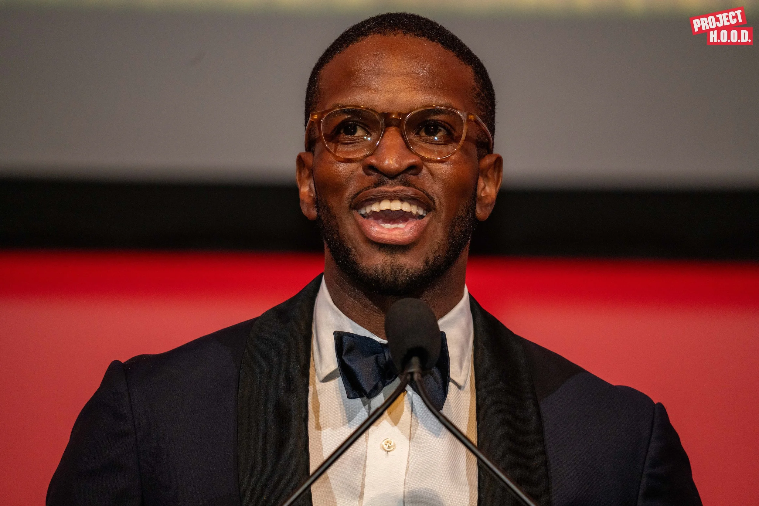 Man wearing glasses, a tuxedo with a bow tie, speaking at a microphone during an event with a red background and the 'Project H.O.O.D.' logo in the top right corner.