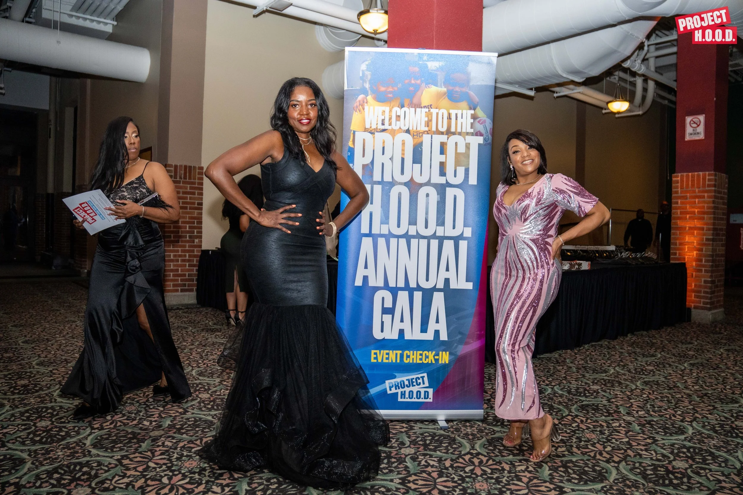 Three women in formal attire standing in front of a large sign that reads 'Welcome to the Project H.O.O.D. Annual Gala'. The woman in the center wears a black mermaid gown, the woman on the right wears a pink and silver sequined gown, and the woman o