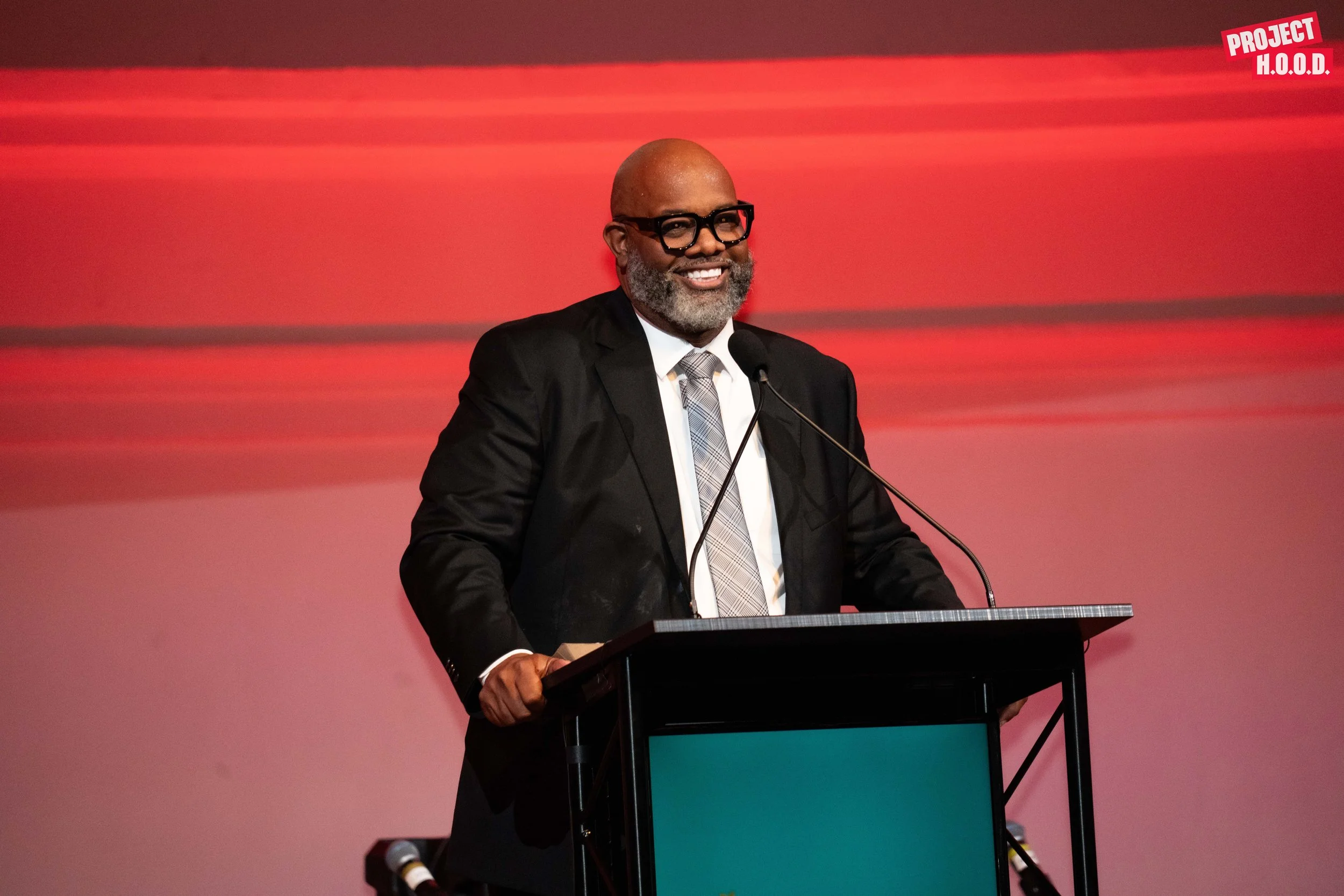 A man in a black suit, white shirt, and plaid tie speaking at a podium with a microphone, smiling, with a red and pink gradient background and a "PROJECT H.O.O.D." logo in the top right corner.