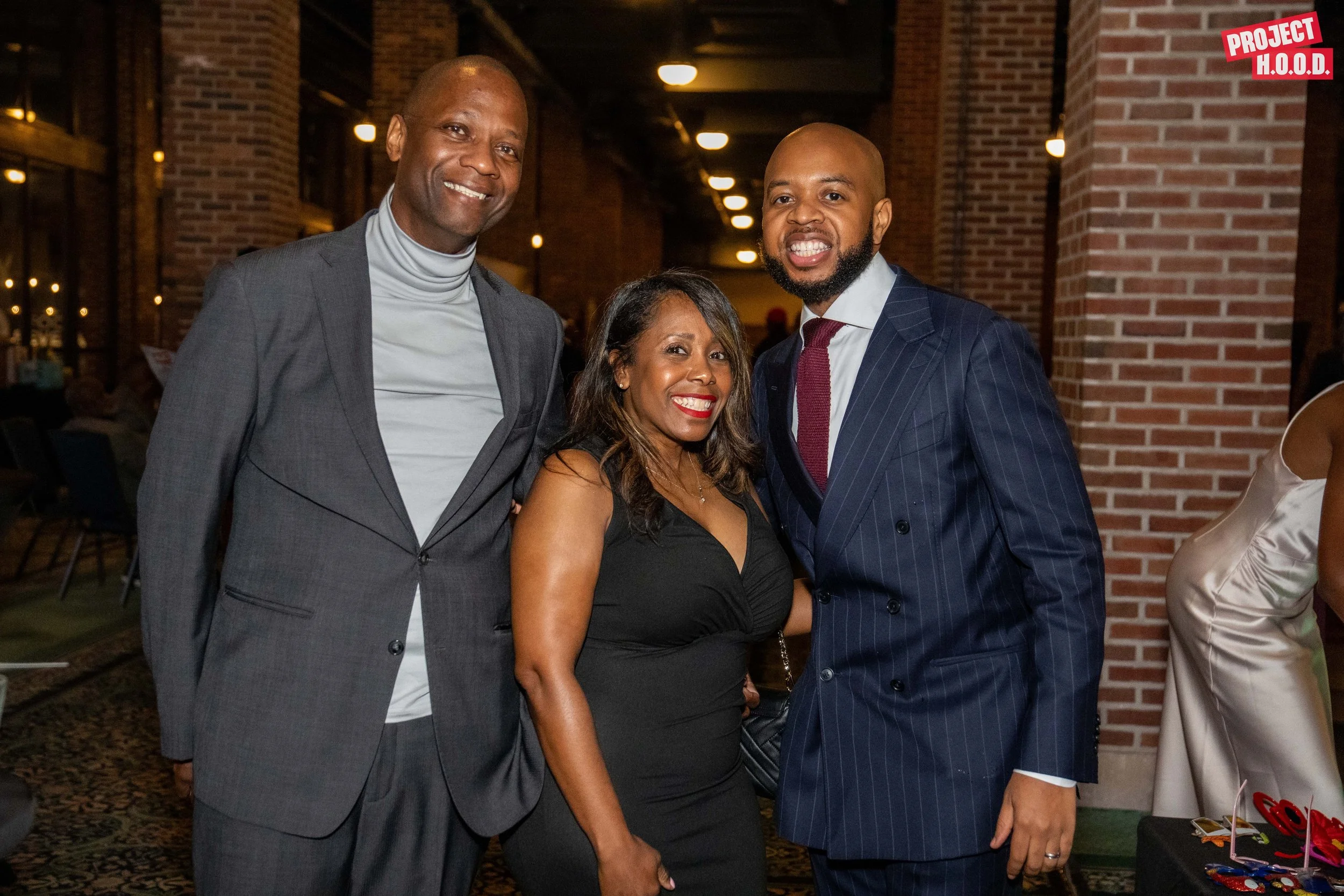 Three people dressed in formal attire smiling at a social event, standing indoors with brick walls and soft lighting.