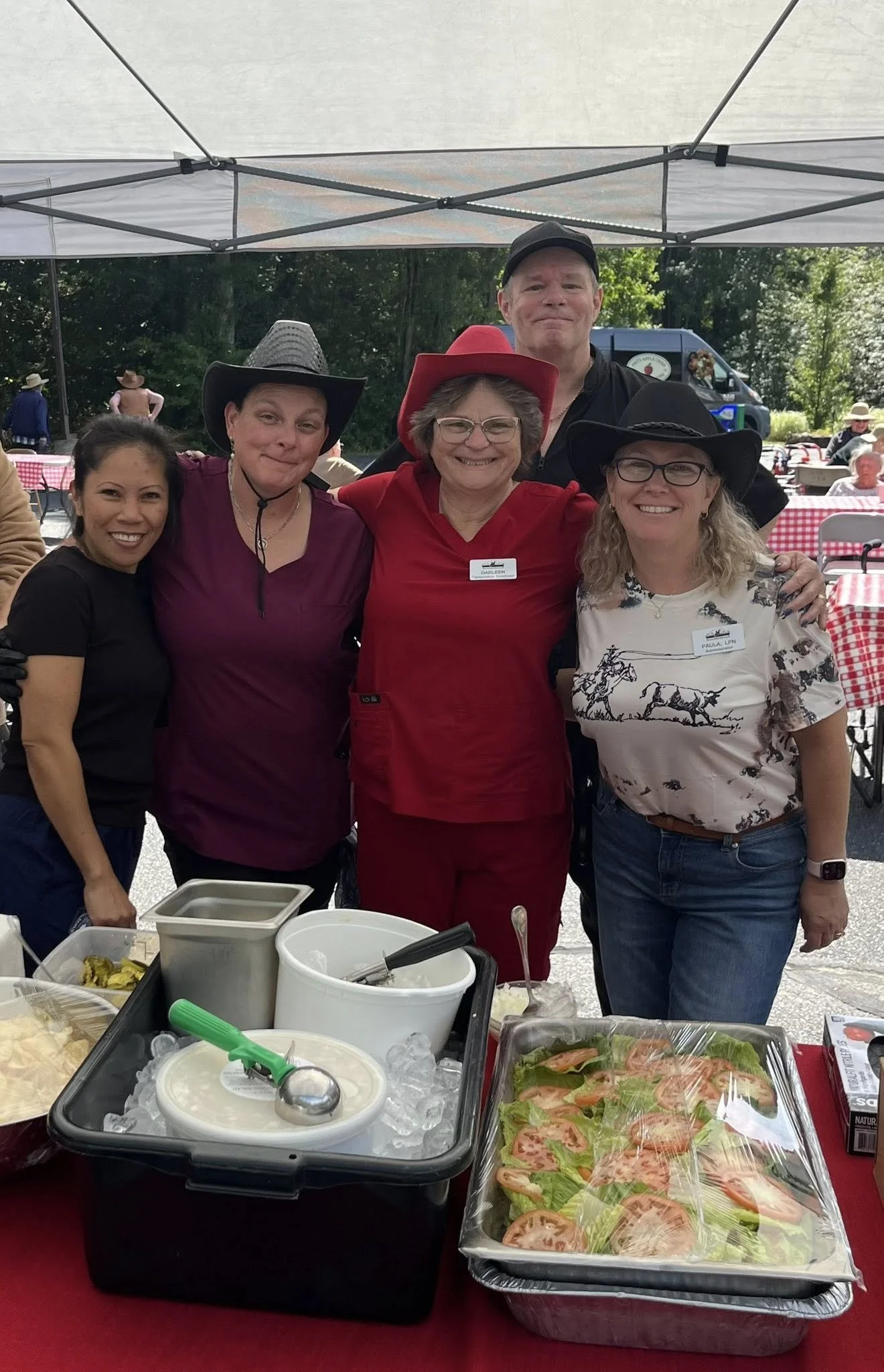 Group of five women and one man smiling at an outdoor event, wearing cowboy hats, with food and drinks on a table in front of them.