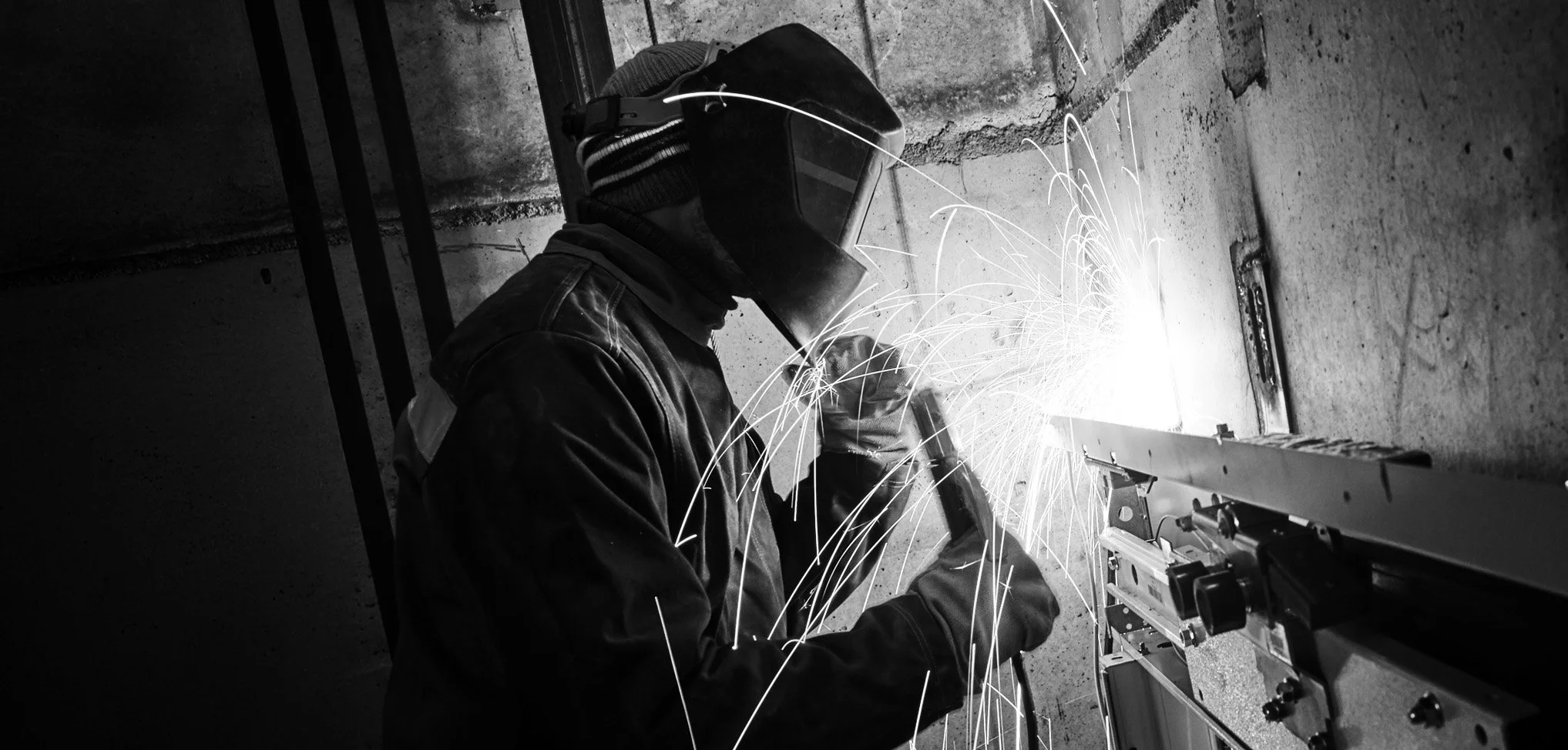 A person welding metal, wearing protective gear including a welding helmet, gloves, and a beanie, with bright sparks flying in a dark industrial setting.