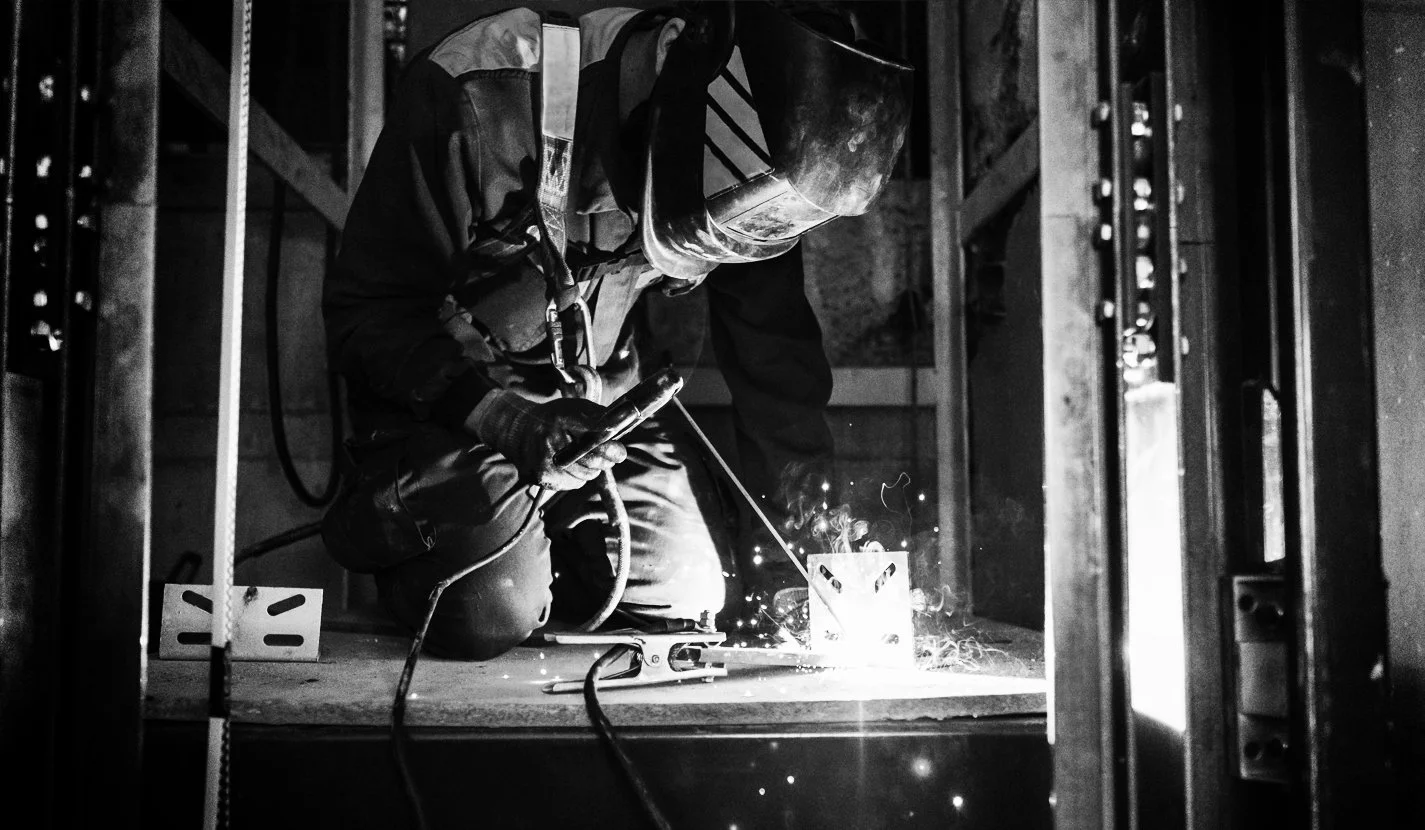 A worker welding, wearing a helmet and gloves, on a construction site with metal framing.