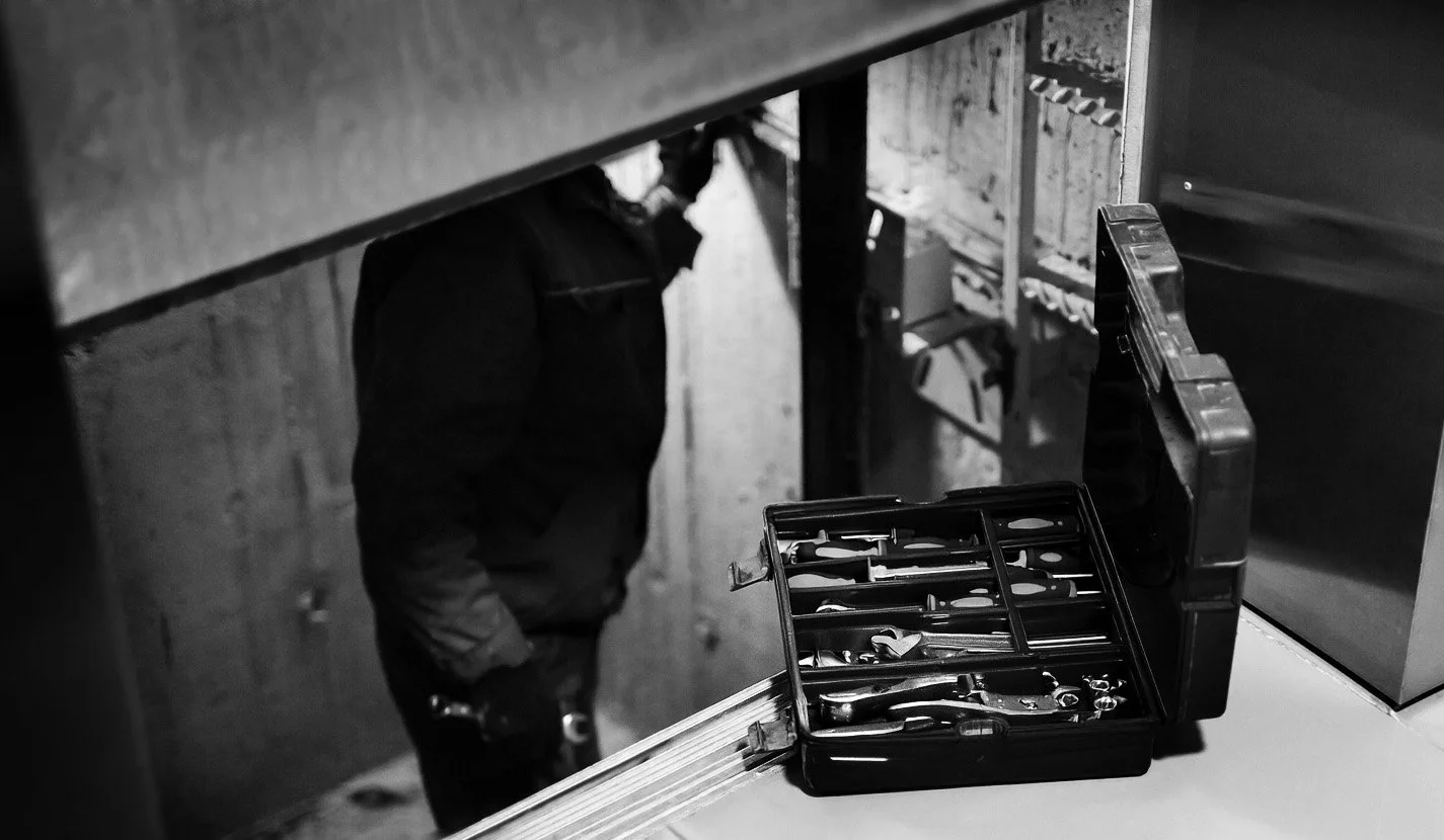 A person wearing dark clothing stands in a dark, industrial-looking environment, with a toolbox of various tools open on a nearby surface.