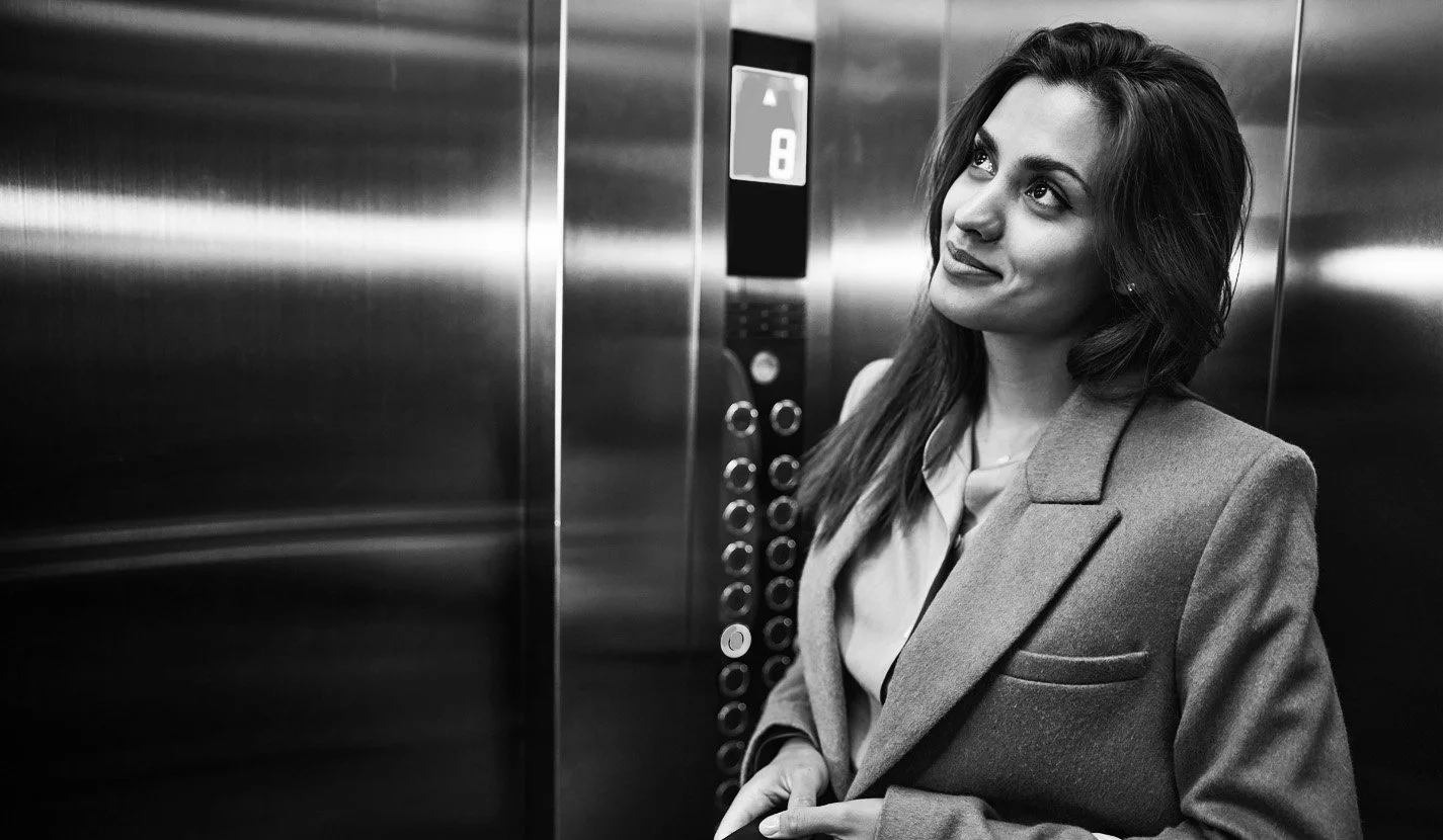 Black and white photo of a woman with long hair, wearing a blazer, standing in an elevator with metallic walls, smiling and looking upwards.