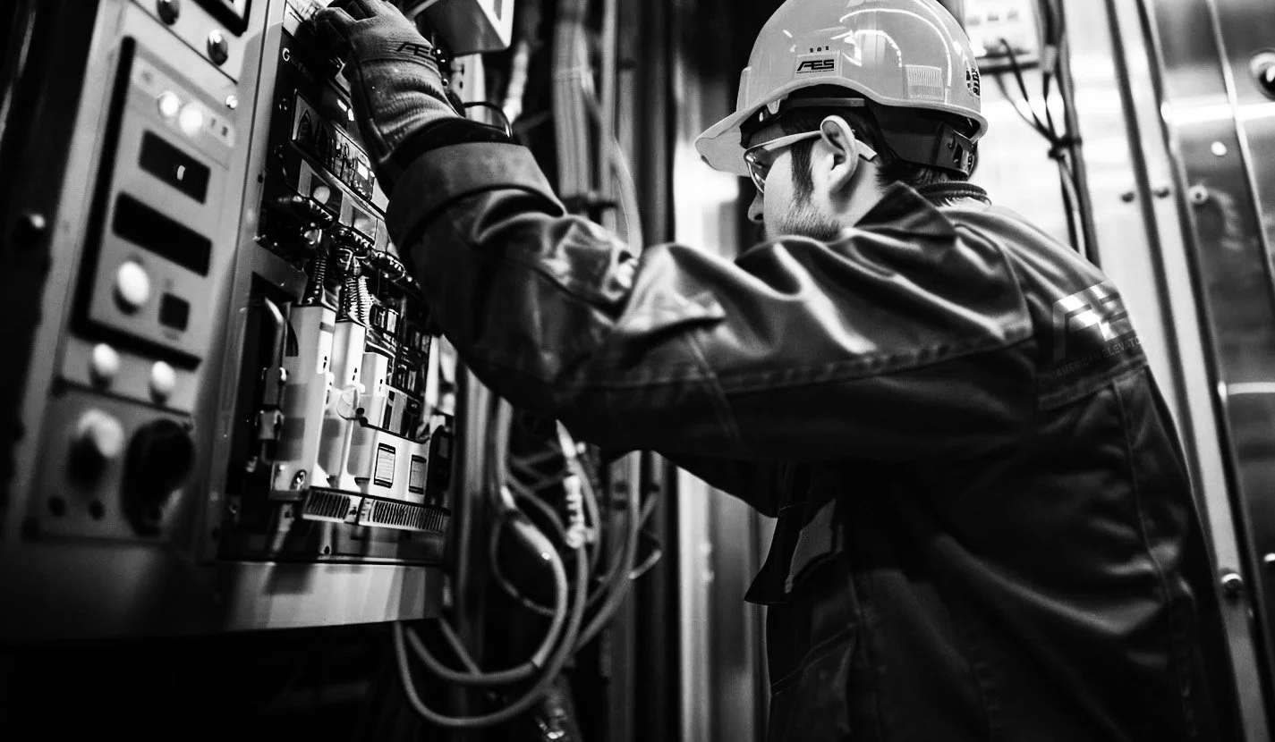 A technician working on electrical equipment inside an industrial facility. The person is wearing a safety helmet, safety glasses, and gloves, focusing on the control panel with various switches and wires.