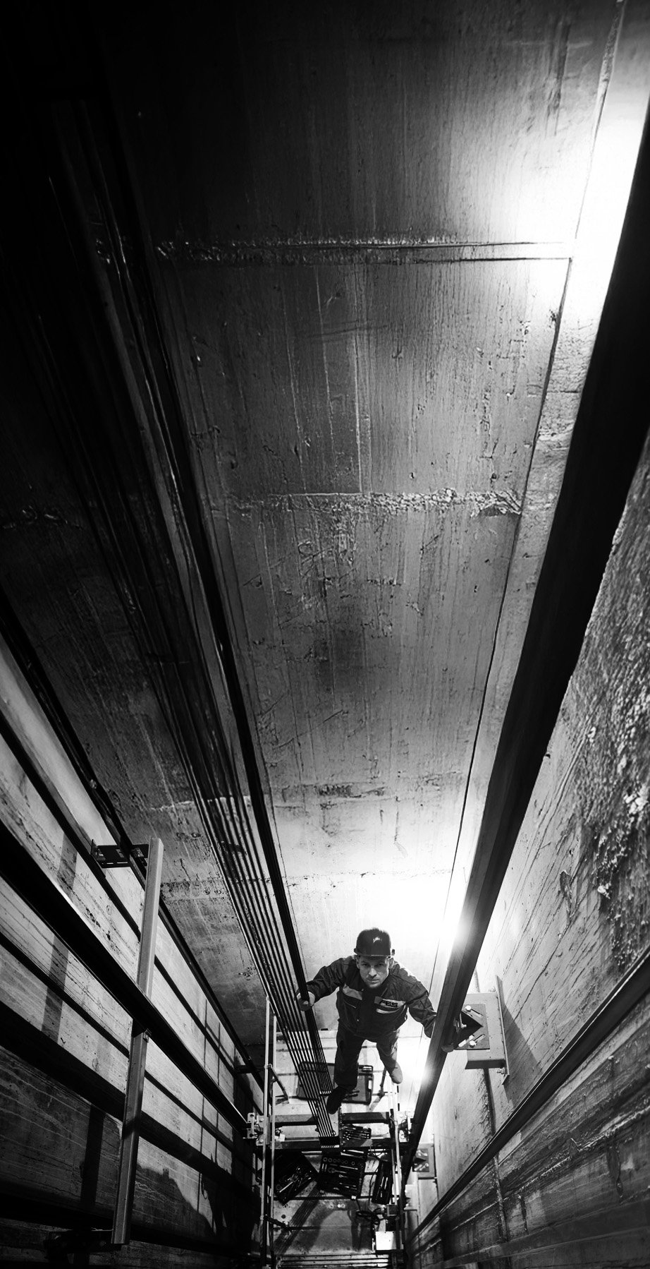 A worker looking up while on a lift inside a deep elevator shaft, with concrete walls and electrical wiring.