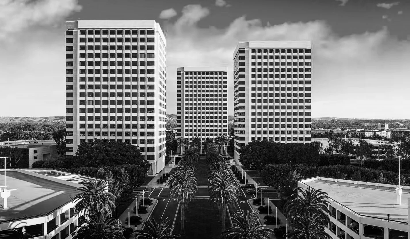 Black and white photo of three tall modern office buildings with a parking lot and palm trees in front, under cloudy sky.