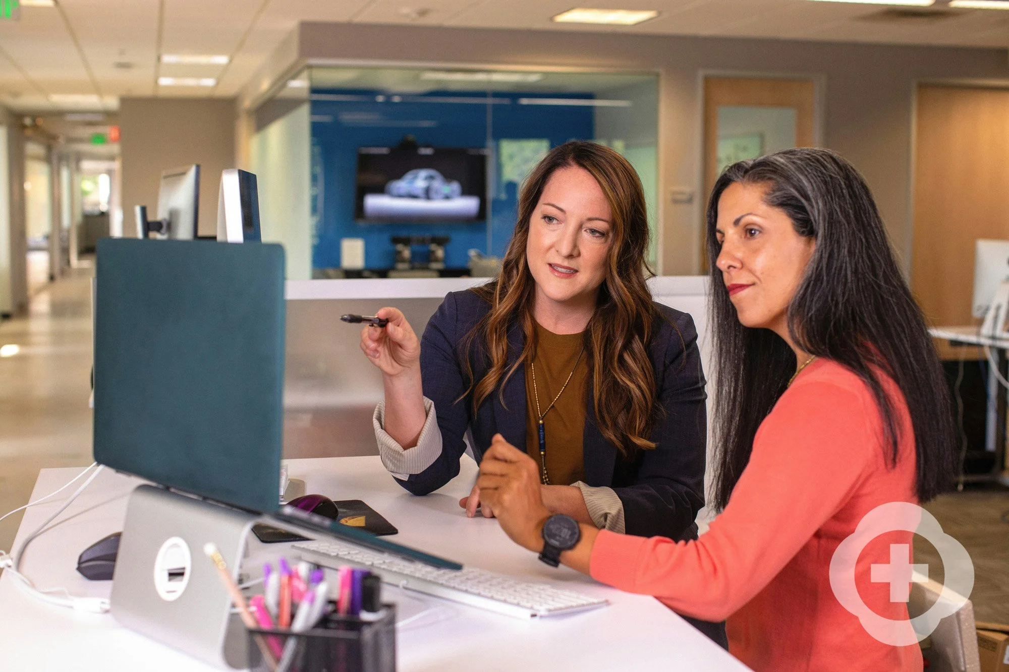 Two women sitting at a desk in an office, engaged in conversation, with a desktop computer and office supplies in the foreground.